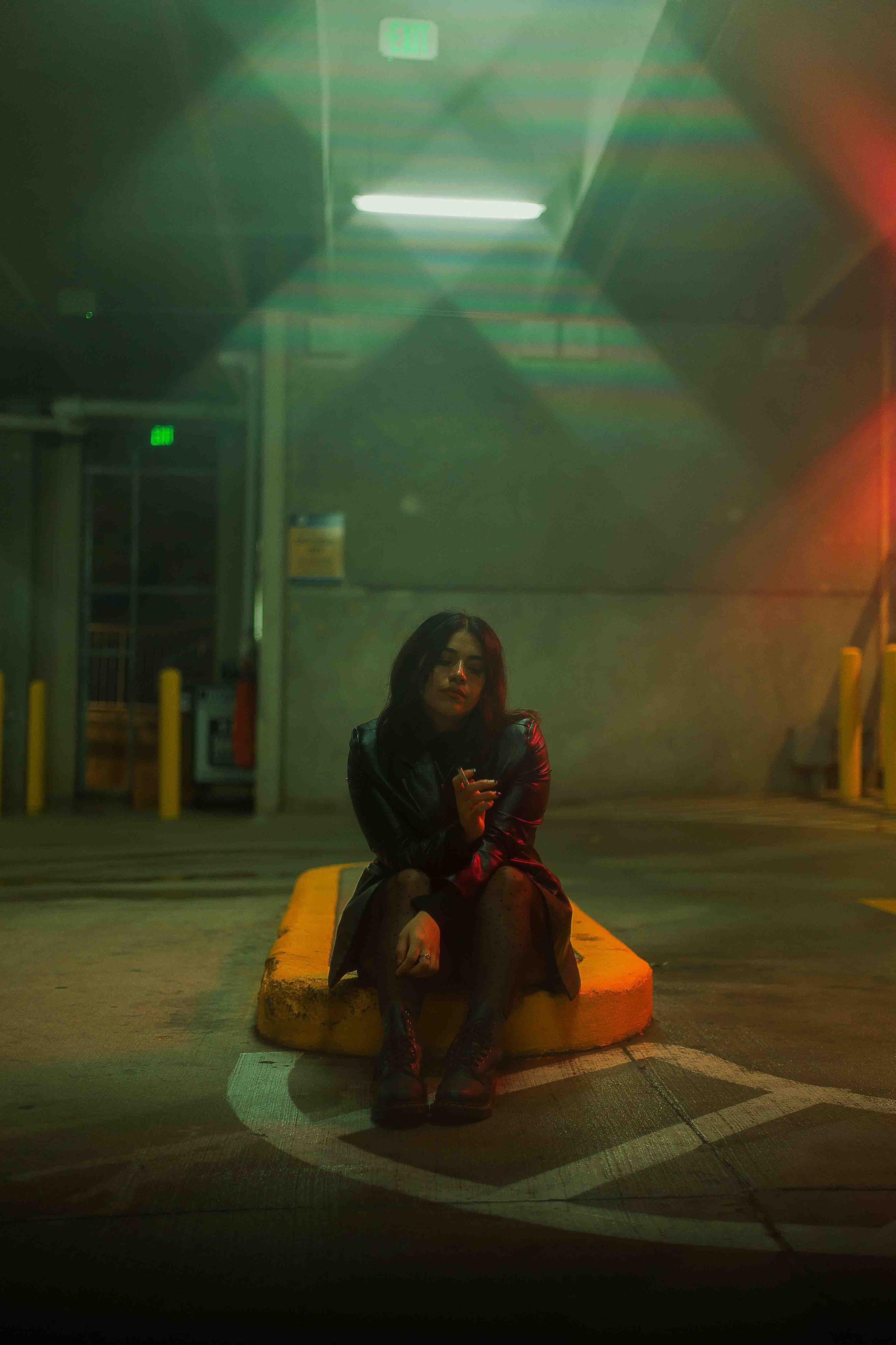 Cinematic night portrait of a woman sitting on a yellow curb in a parking lot wearing black boots, a black leather jacket, and a patterned dress with neon lighting casting colorful reflections