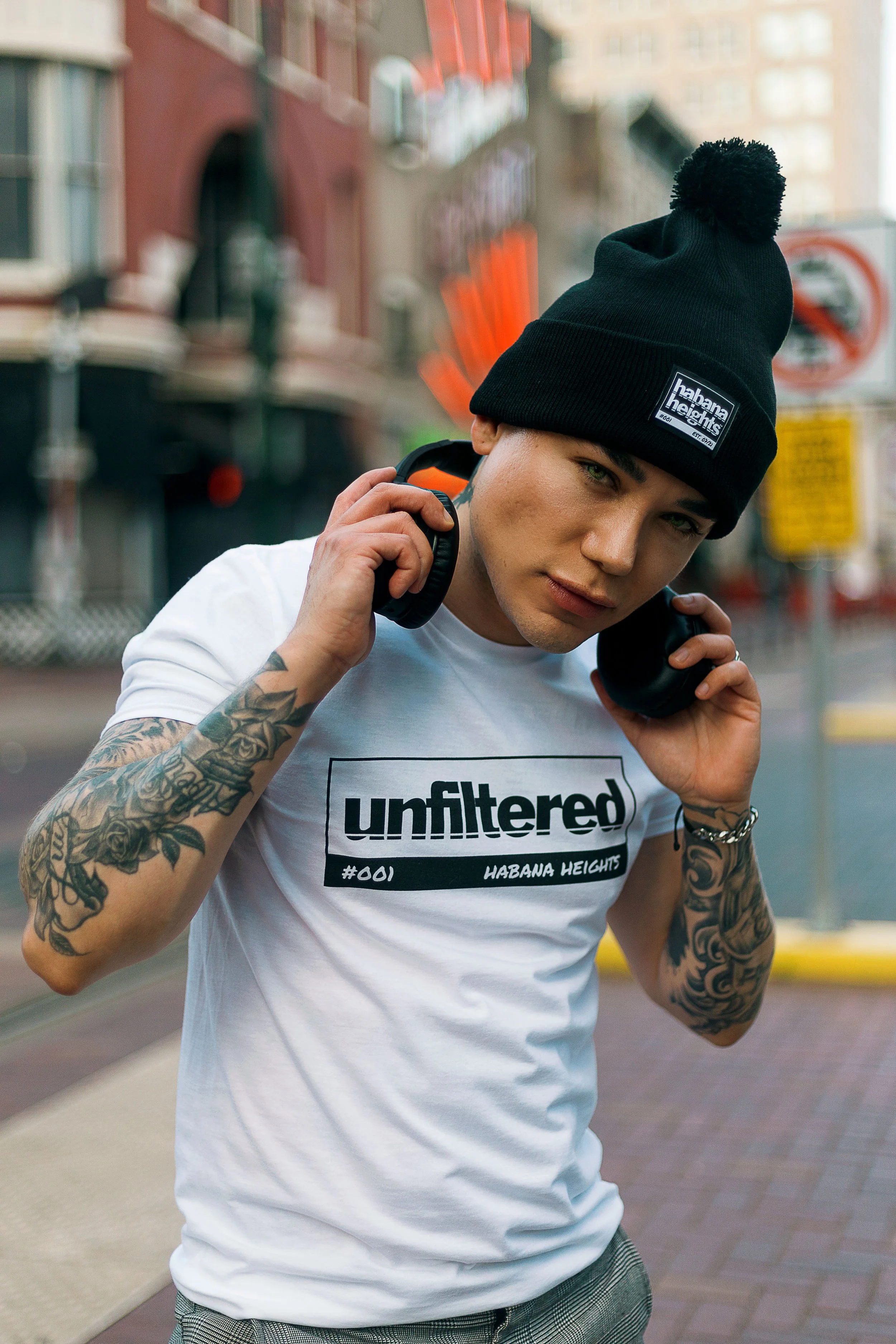 Young man wearing a black beanie with a patch, white t-shirt with bold black text, tattoos on both arms, holding headphones around his neck, standing on city street.