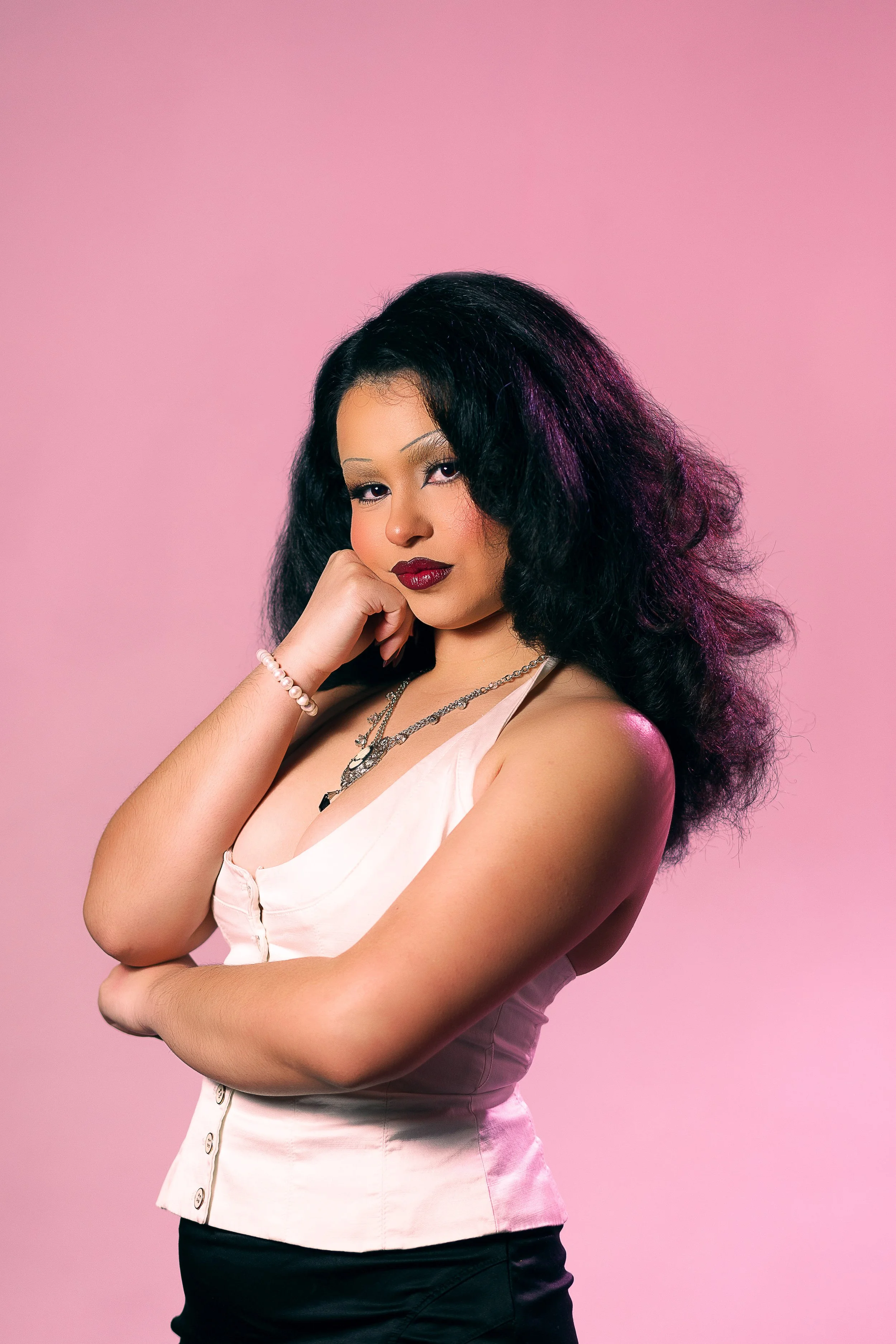 A woman with voluminous black hair with purple highlights, wearing a sleeveless white top, jewelry, and dark lipstick, is posing with one hand near her face against a pink background.