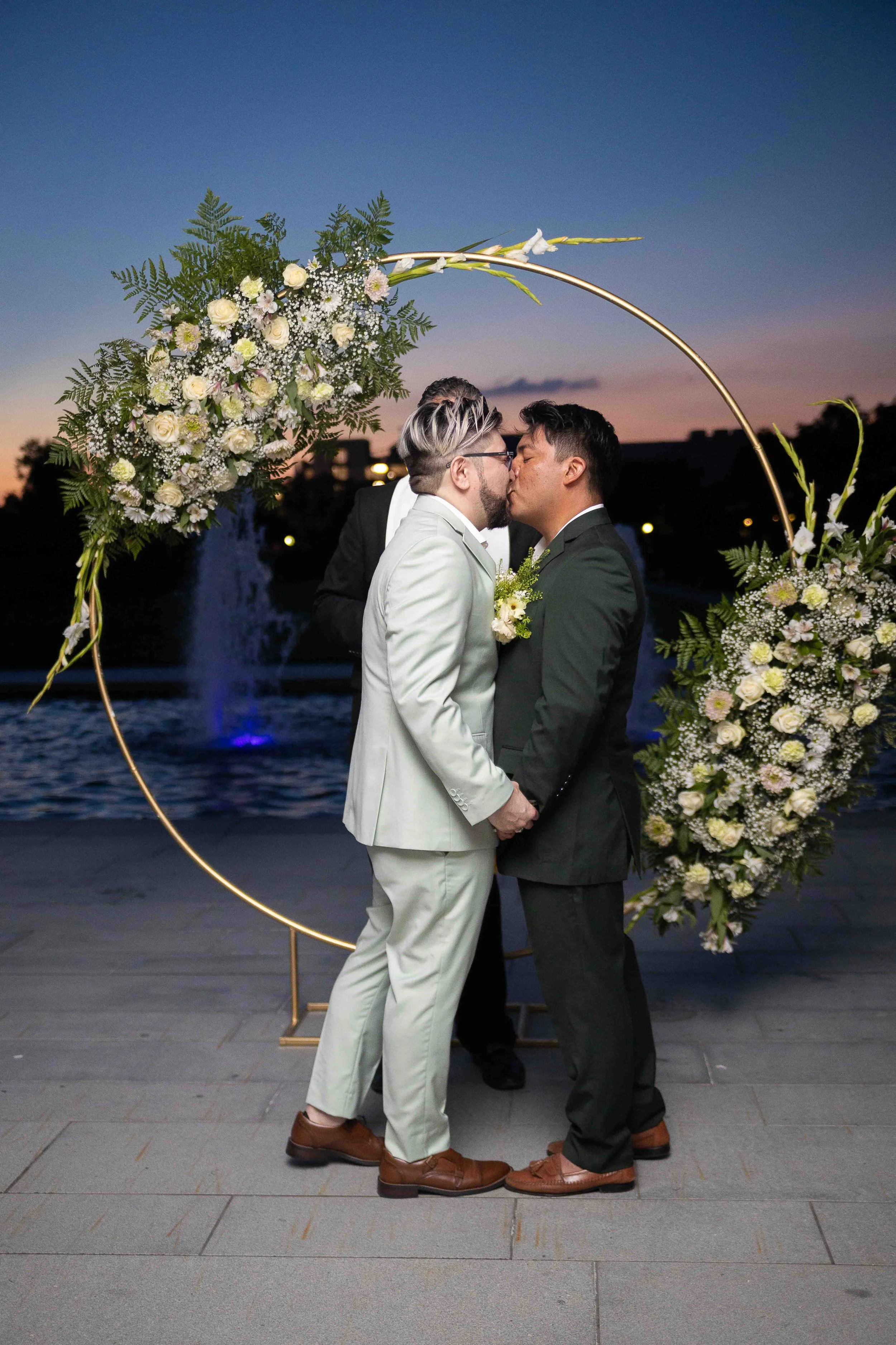  LGBTQ+ wedding ceremony at McGovern Centennial Gardens in Houston, Texas by Lev’s Photography featuring a same-sex couple kissing at sunset in front of a circular floral arch with white and pink flowers near a water fountain