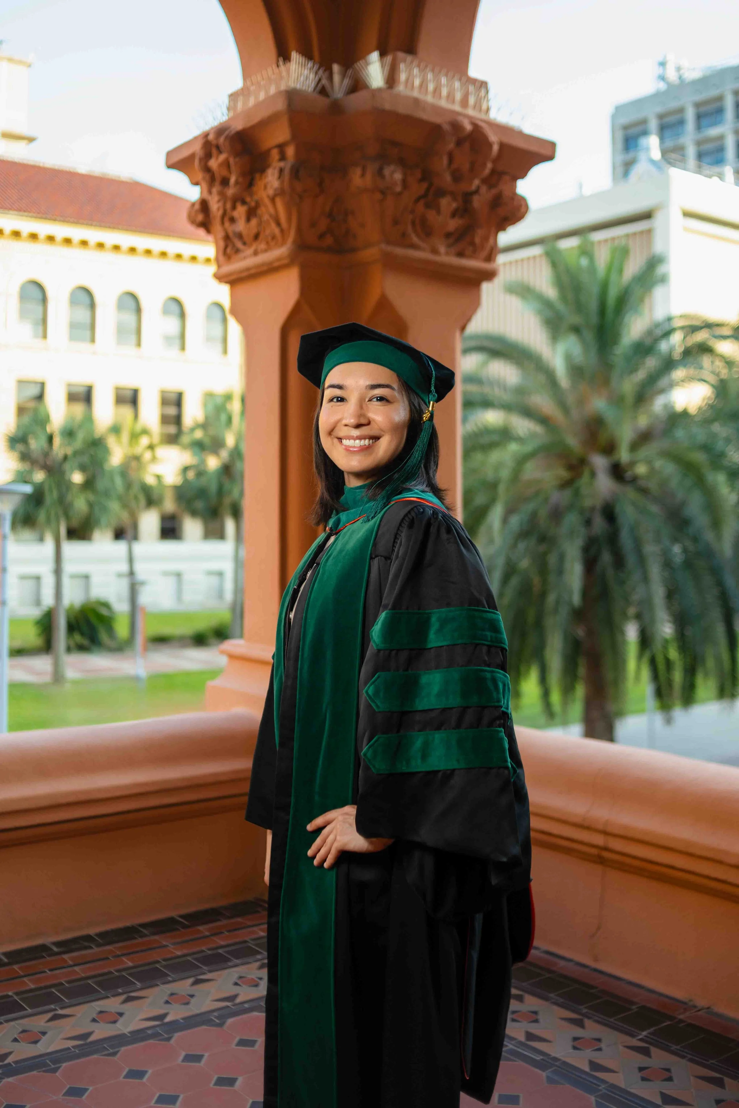 Graduation portrait at Old Red in Galveston, Texas by Lev’s Photography featuring a smiling woman in cap and gown standing on campus with palm trees and historic buildings in the background