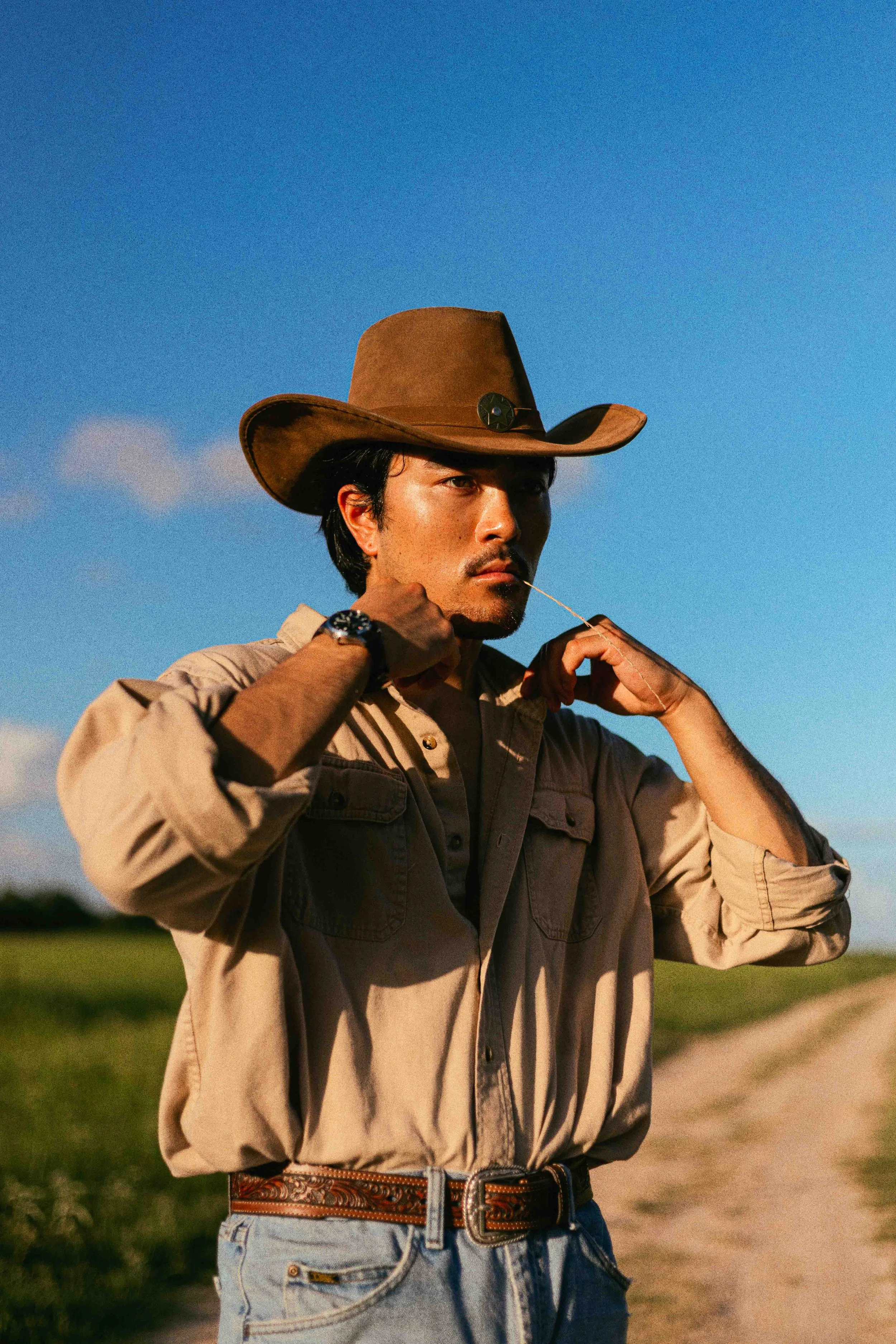 Western cowboy portrait of a man in tan shirt and cowboy hat standing outdoors on a dirt path under blue sky in Houston, Texas