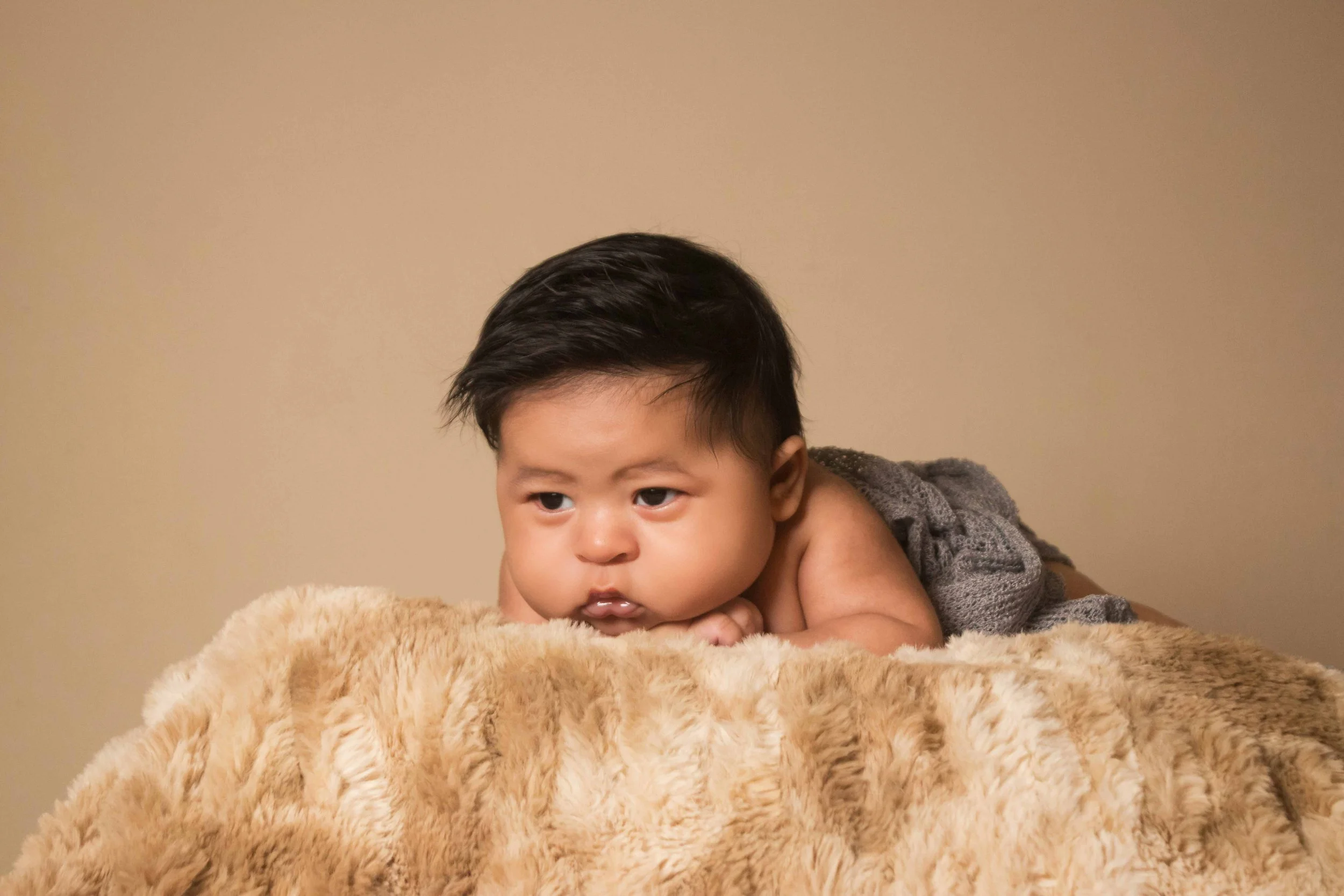 Newborn portrait in Houston, Texas by Lev’s Photography featuring a young Mexican baby lying on a soft beige blanket, resting his chin on his hands, and looking thoughtfully to the side against a plain beige background