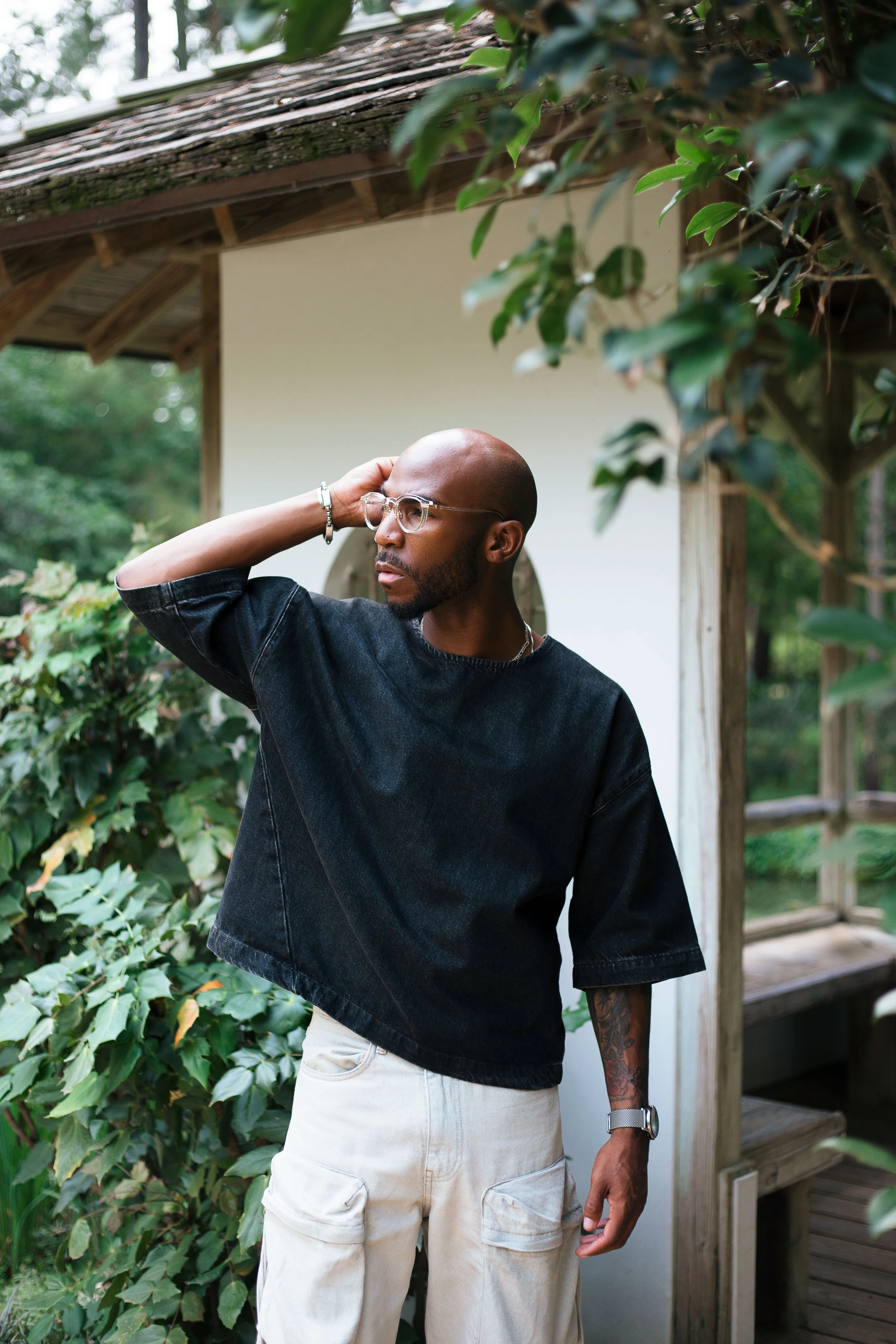A bald man with glasses, wearing a black oversized t-shirt and beige cargo shorts, standing outdoors near a wooden structure and surrounded by green foliage, with his hand behind his head, looking to the side.