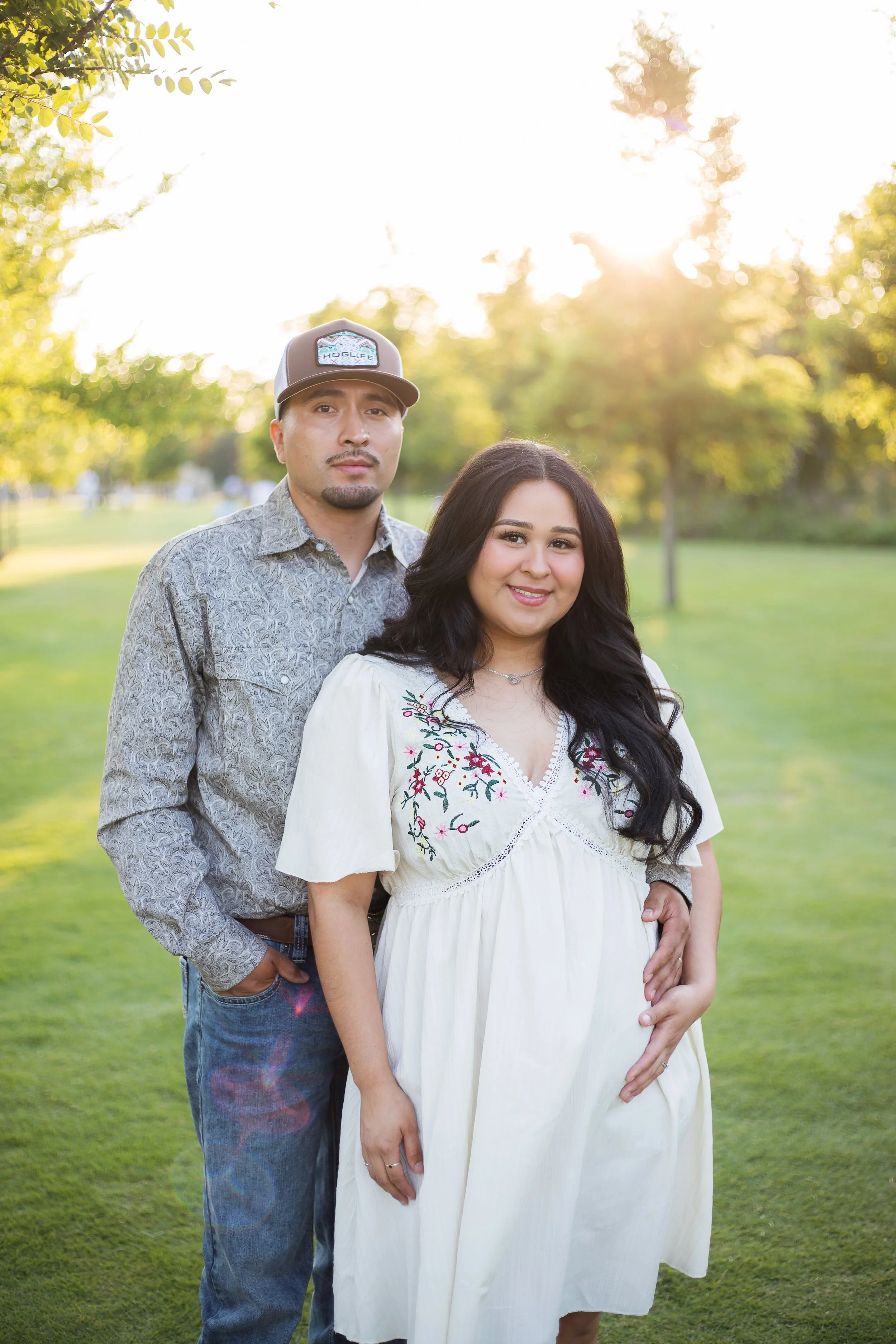 A pregnant woman and a man standing outdoors on grassy ground with trees in the background, illuminated by the setting sun.