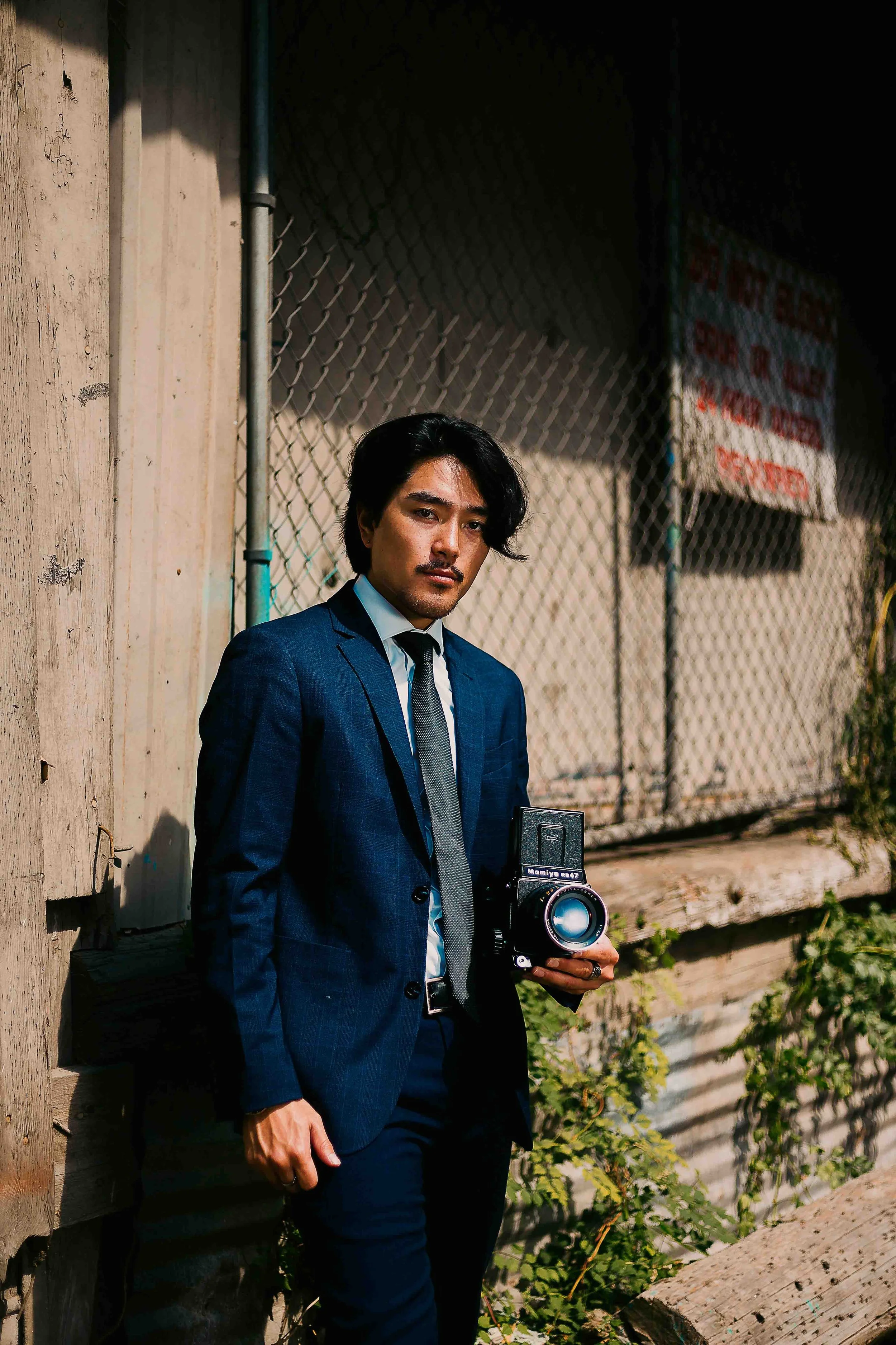 Professional portrait session in Houston, Texas by Lev’s Photography featuring a man in a dark blue suit holding a Mamiya RB67 camera outdoors near a wooden wall with dramatic sunlight and shadows.