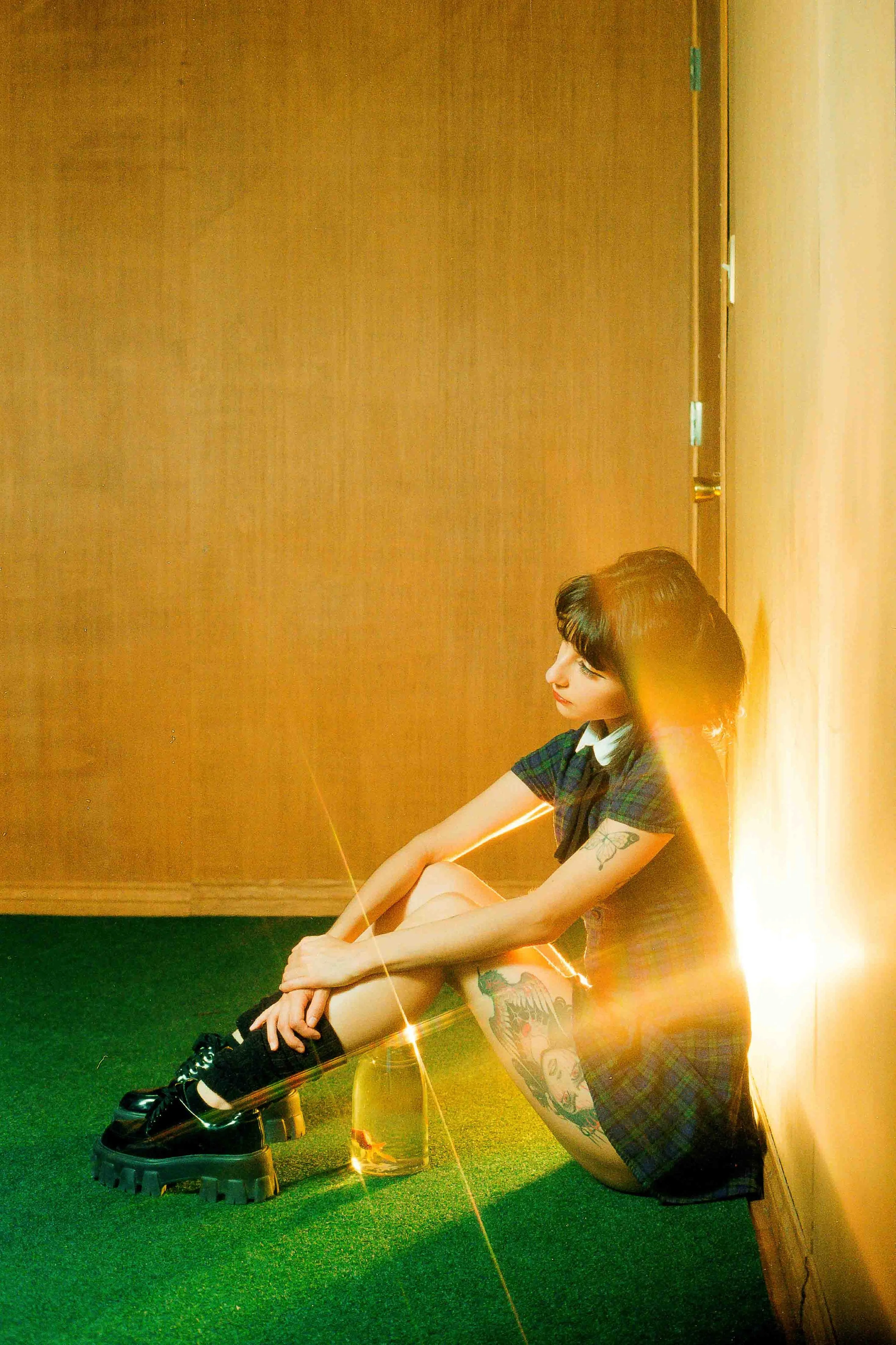 Editorial portrait of a tattooed woman sitting on a carpeted floor, leaning against a wall with her knees drawn up and arms wrapped around her legs, wearing a plaid dress and black boots, illuminated by bright light with lens flare in a creative colo