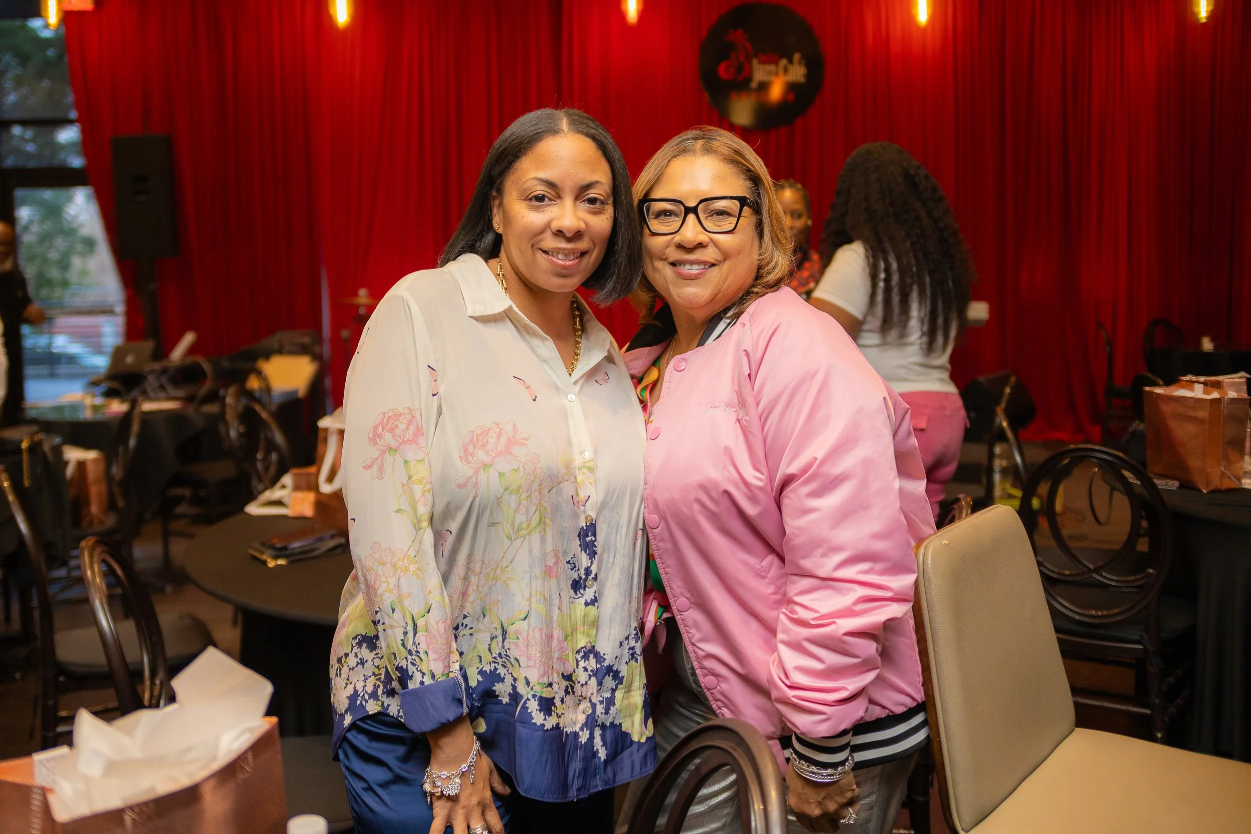 Two women smiling at the camera at a social gathering. One is wearing a floral blouse, and the other is wearing a pink jacket. They are standing in a room with red curtains and tables around them.
