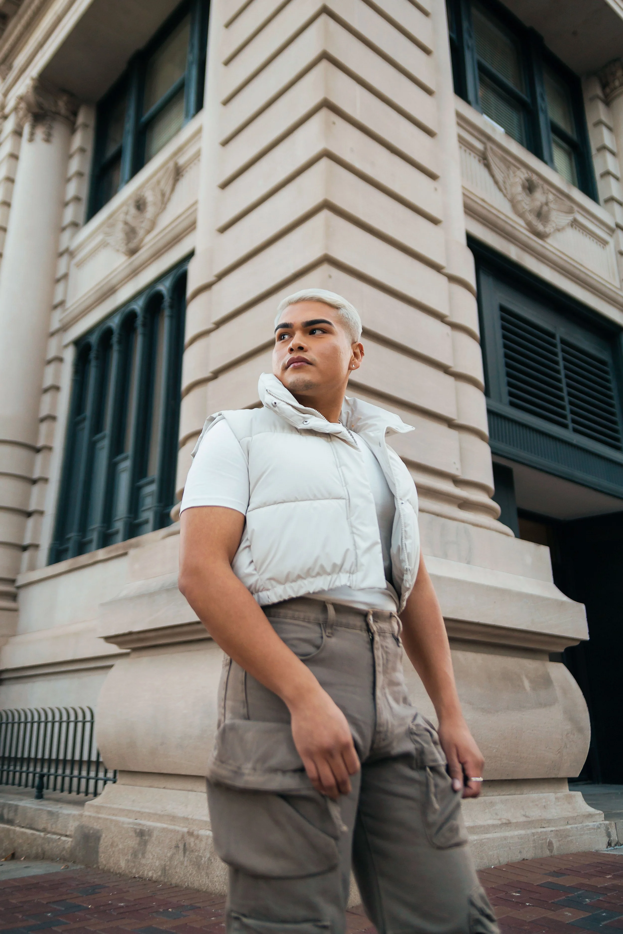 A young person with platinum blonde hair, wearing a white vest over a white t-shirt and beige cargo pants, stands in front of an ornate stone building with large windows and decorative carvings.