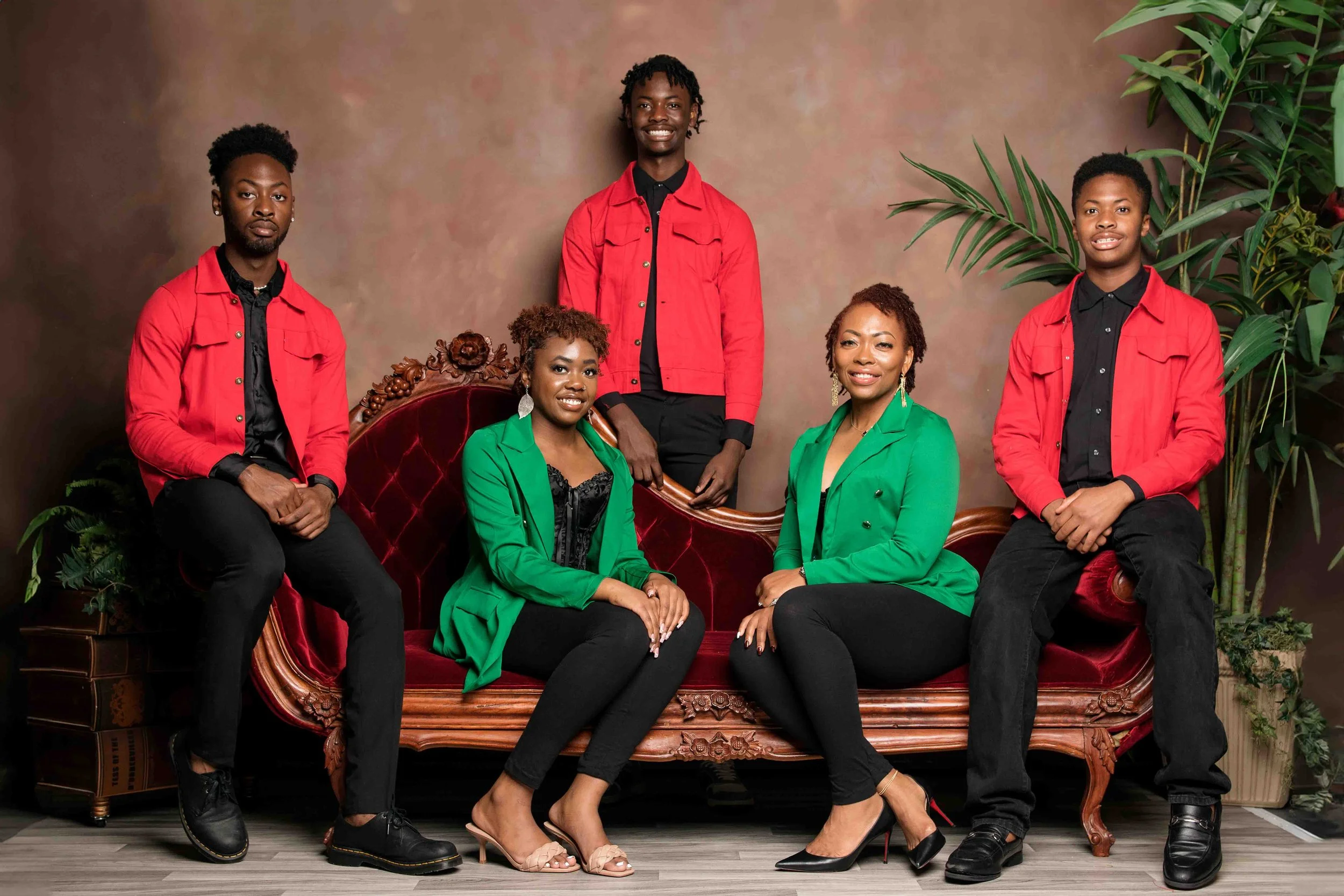 Studio family portrait in Missouri City, Texas by Lev’s Photography featuring six individuals — three women seated on a red velvet sofa and three men standing behind — wearing coordinated fashion outfits with green blazers and red jackets against a b