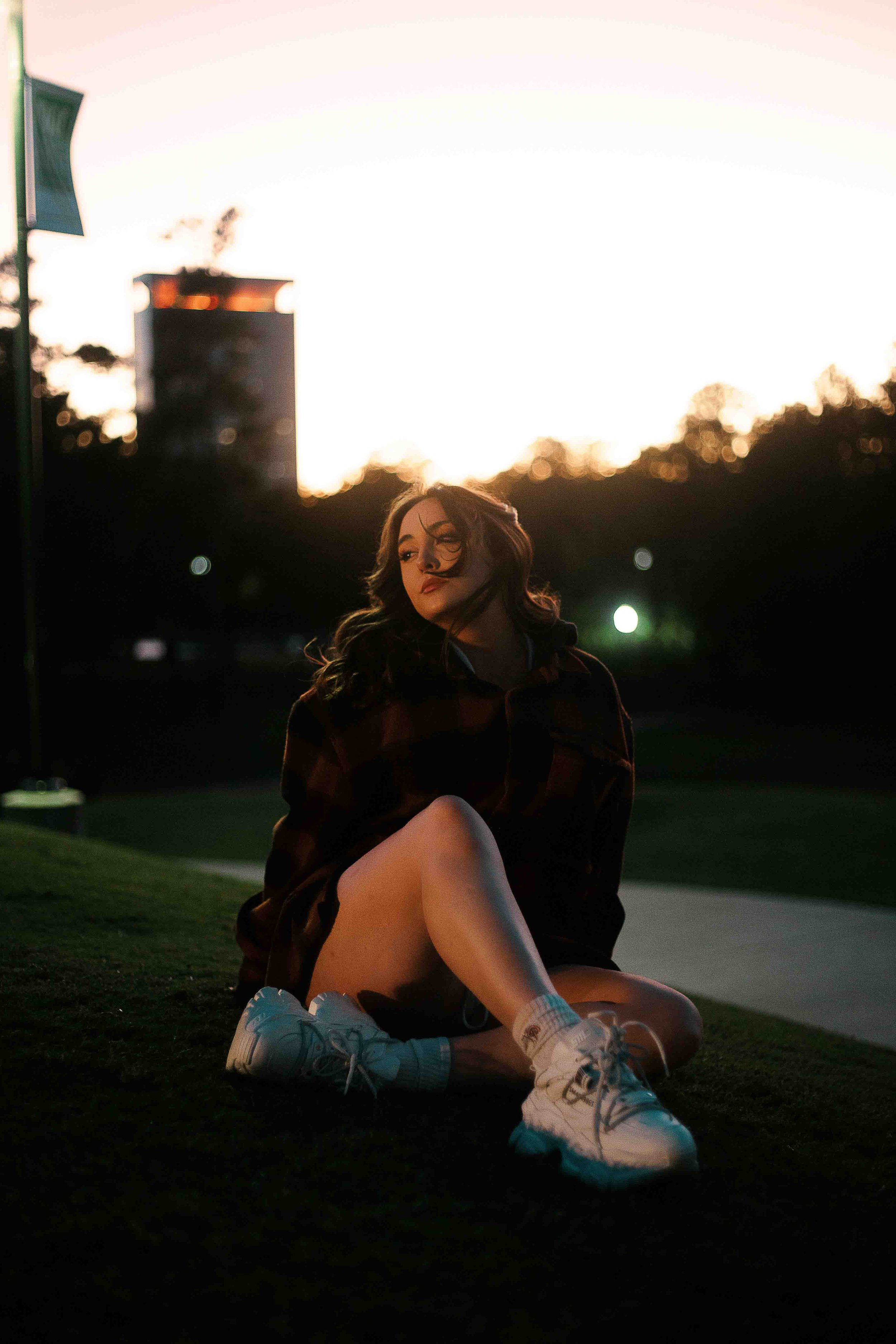 Sunset portrait of a young woman sitting on grass wearing white sneakers, shorts, and a dark jacket with wind blowing through her hair