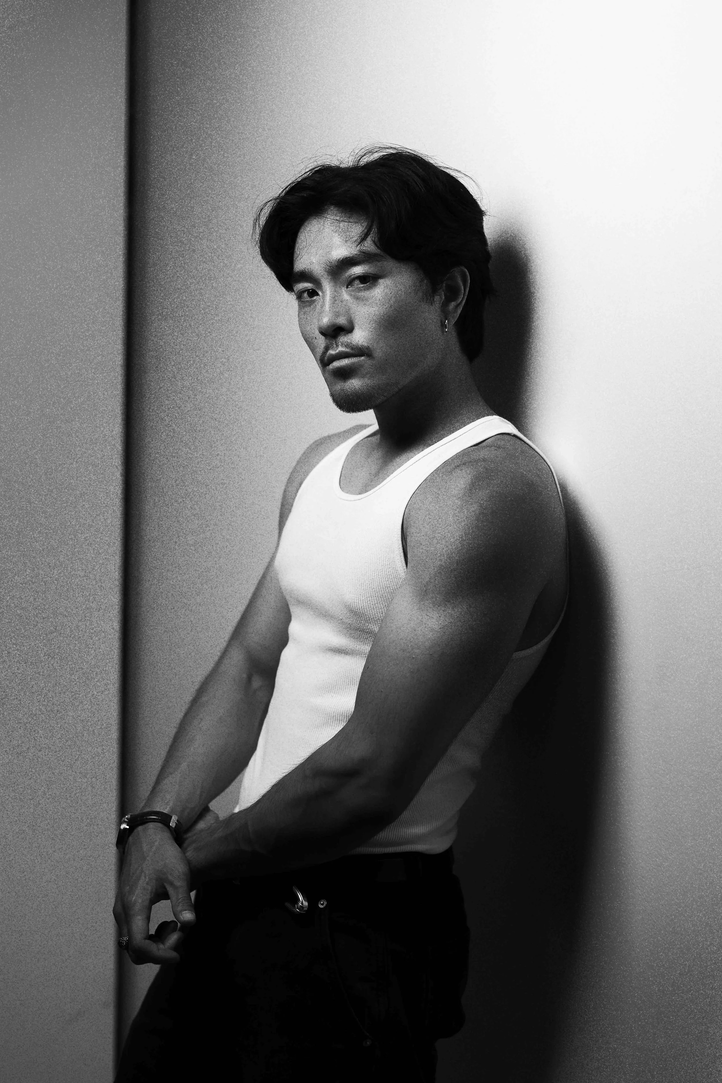 Black and white editorial portrait in Houston, Texas by Lev’s Photography featuring a man with dark hair in a sleeveless white shirt leaning against a wall with a serious expression and grainy texture