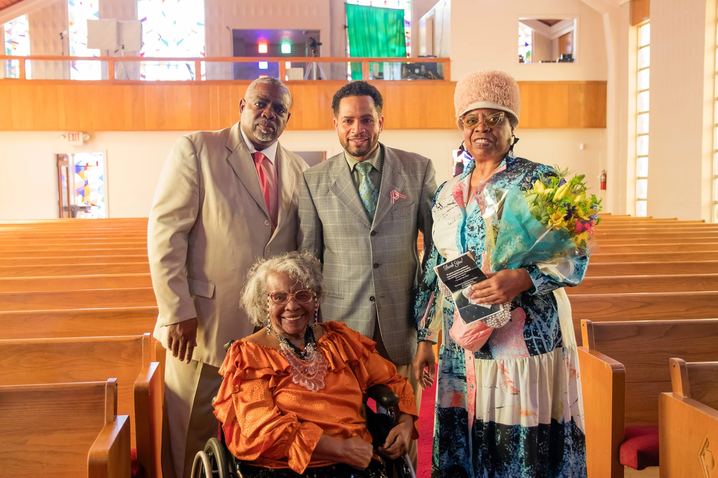 A diverse group of five people, including a woman in a wheelchair, celebrating inside a church with wooden pews and stained glass windows, some holding flowers and a small card.