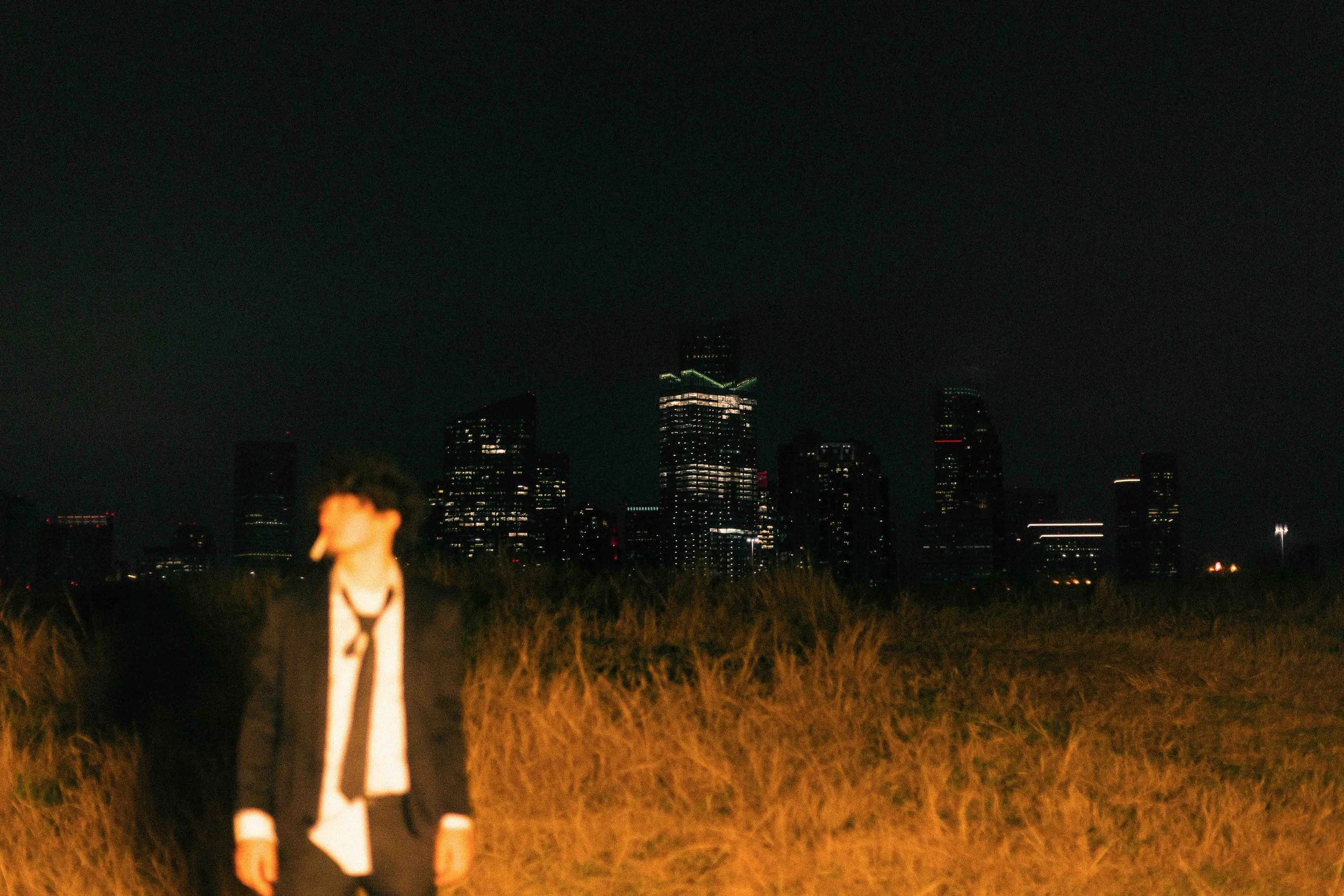 Houston editorial portrait of a person standing in a field of tall grass at night with city skyline and illuminated skyscrapers in the background