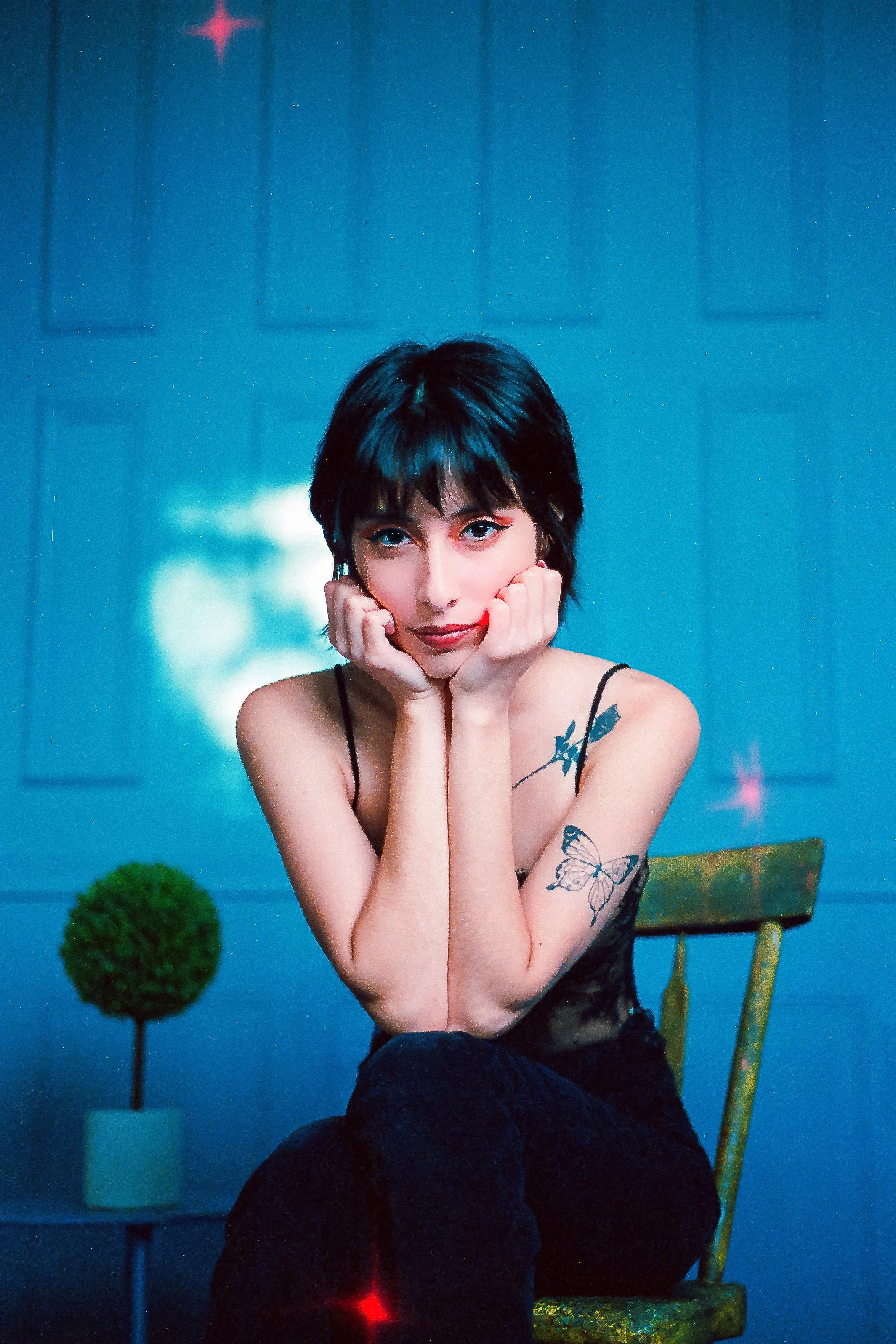 A woman with short black hair and tattoos on her arms, sitting on a wooden chair with her hands resting on her chin, in front of a blue wall with soft lighting.