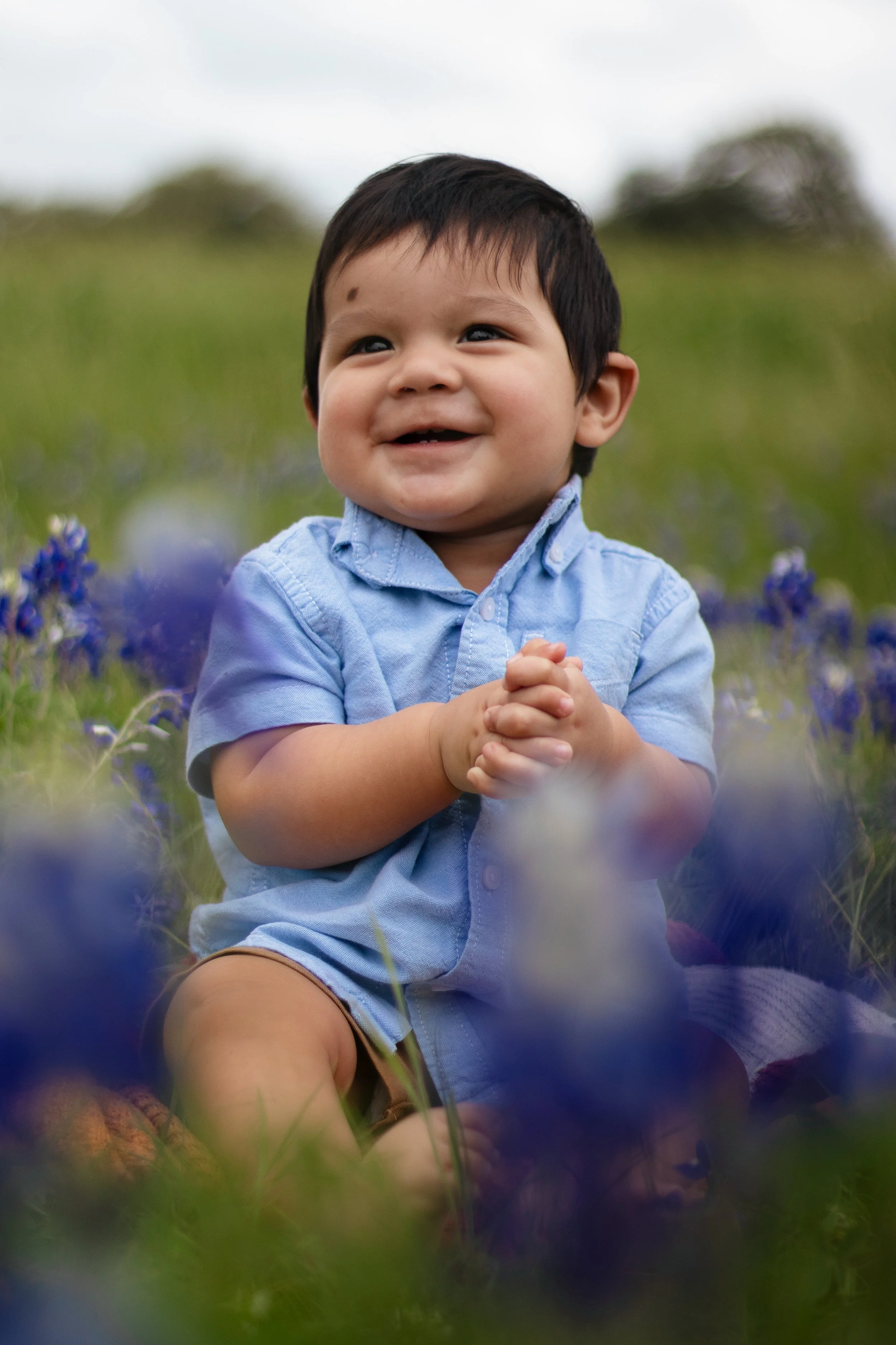 A smiling young boy sitting outdoors in a field of purple flowers, wearing a light blue shirt.