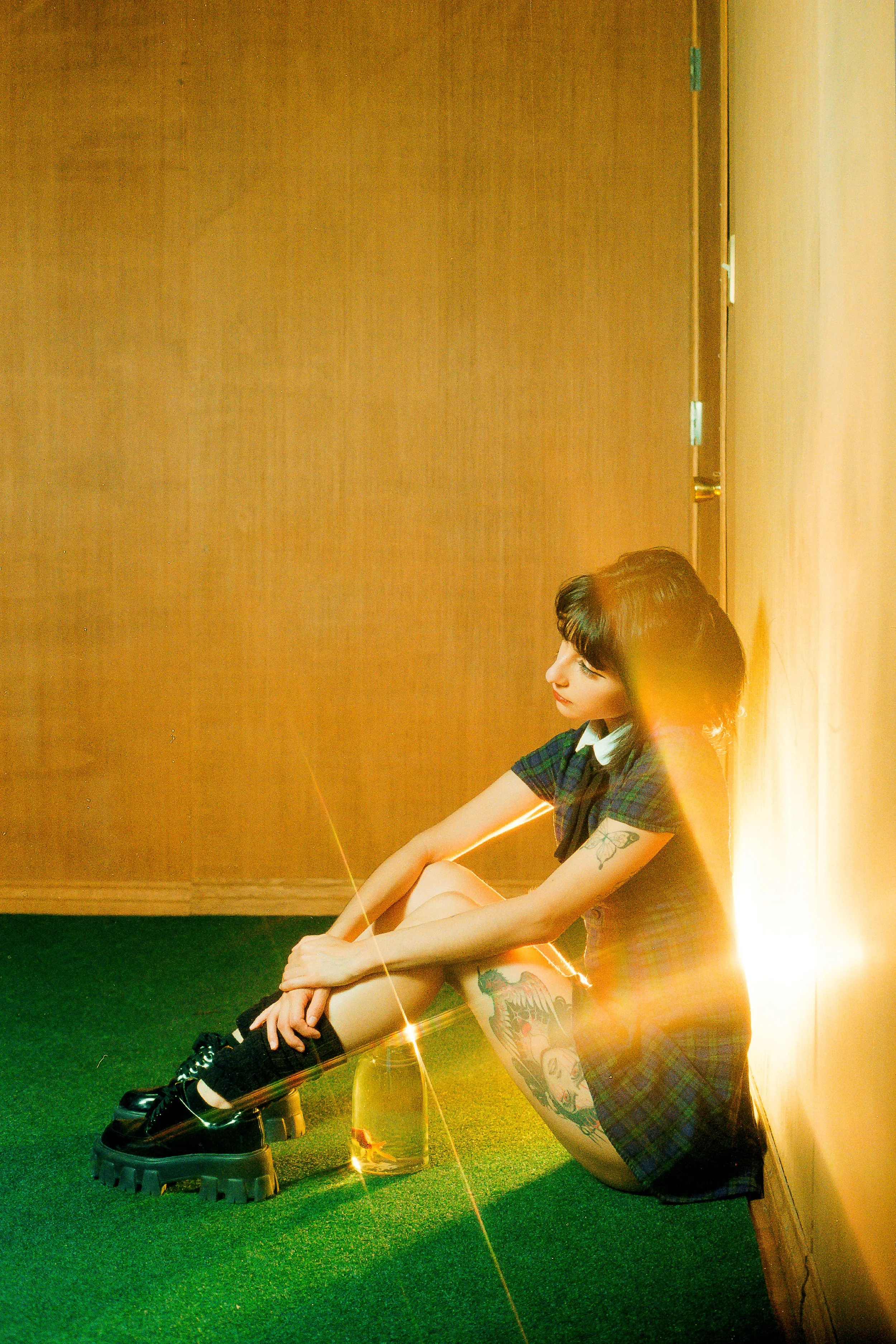 A woman with tattoos sitting on the floor in a room with wooden walls and green carpet, illuminated by yellow sunlight and lens flare.