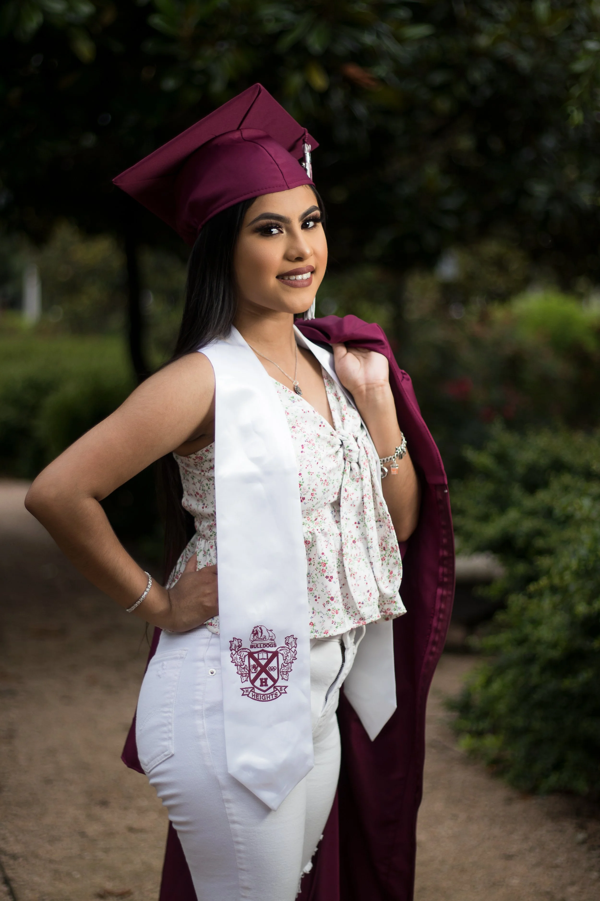 Young woman in a maroon graduation cap and gown, holding the gown over her shoulder, standing outdoors with greenery in the background.