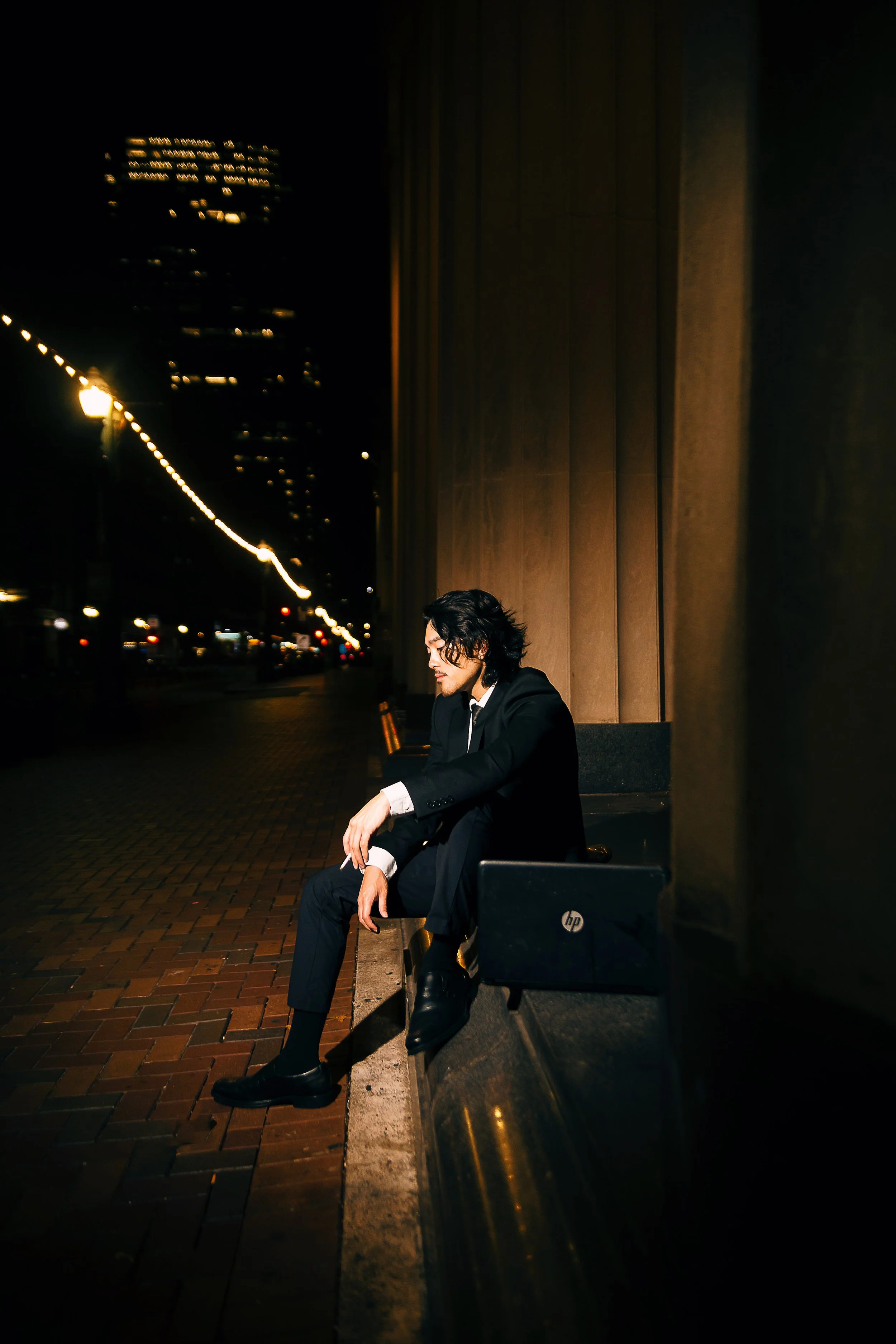 Man in a black suit sitting alone on a bench at night, with a laptop beside him, looking down thoughtfully.