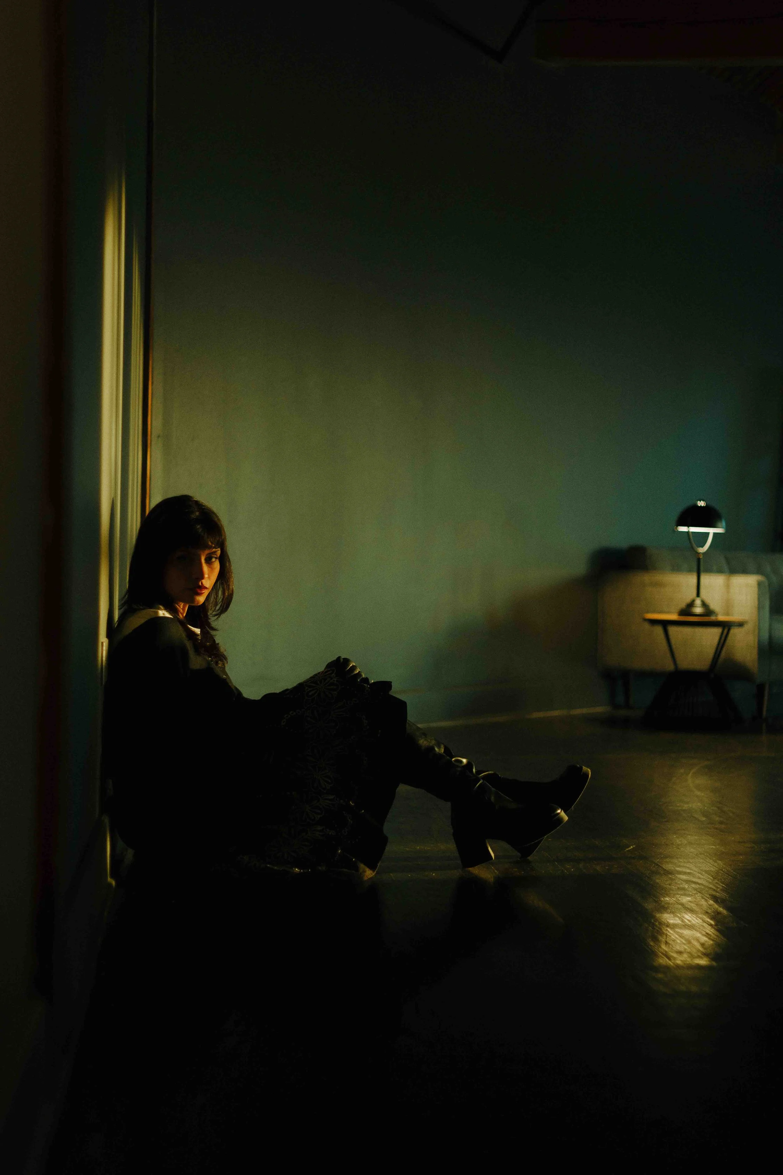 Editorial creative portrait of a woman with dark hair sitting on the floor in a dimly lit room with a sofa and lamp in the background