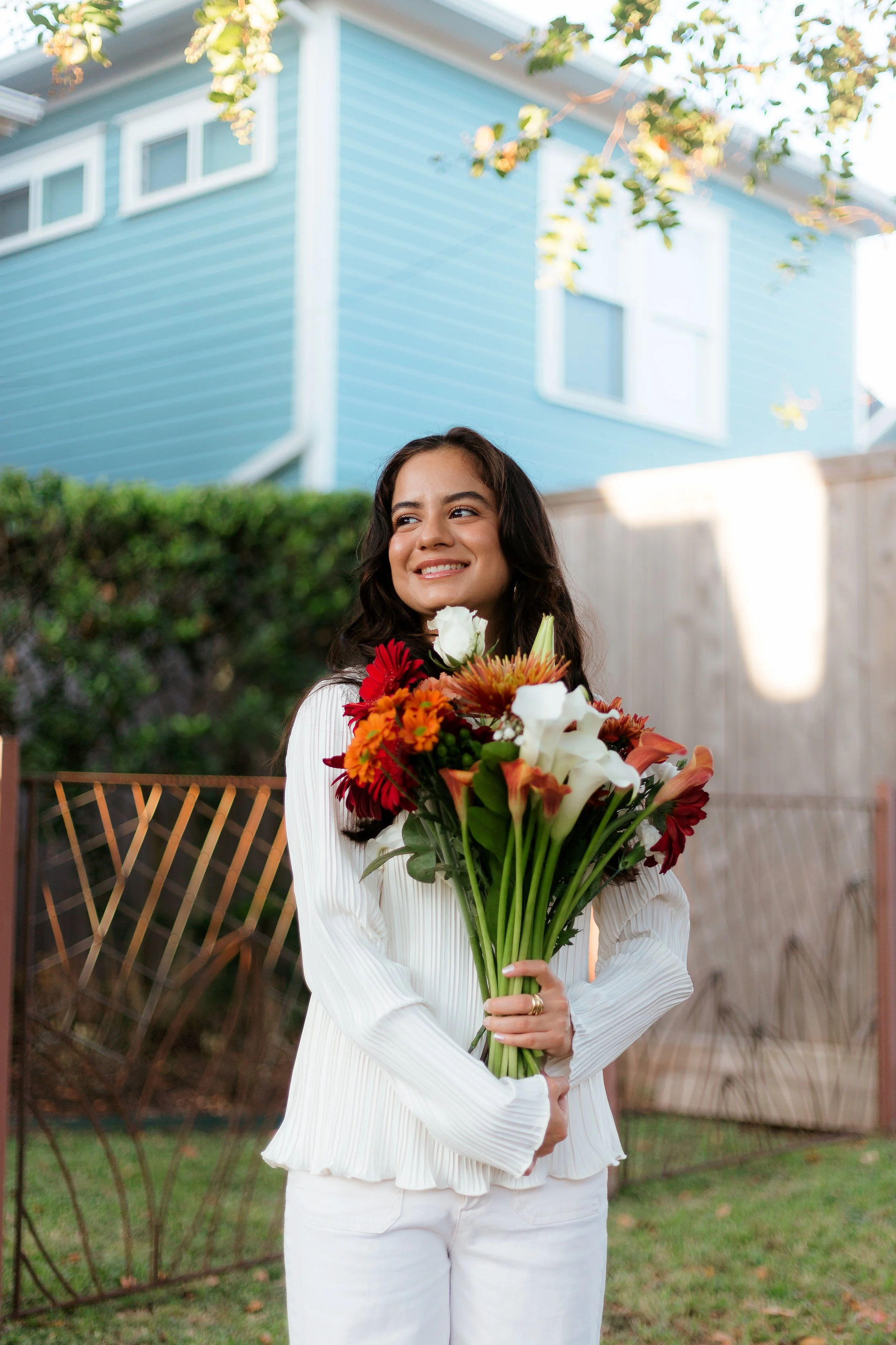 A woman smiling and holding a large bouquet of colorful flowers outdoors, in front of a backyard with a wooden fence and a blue house.