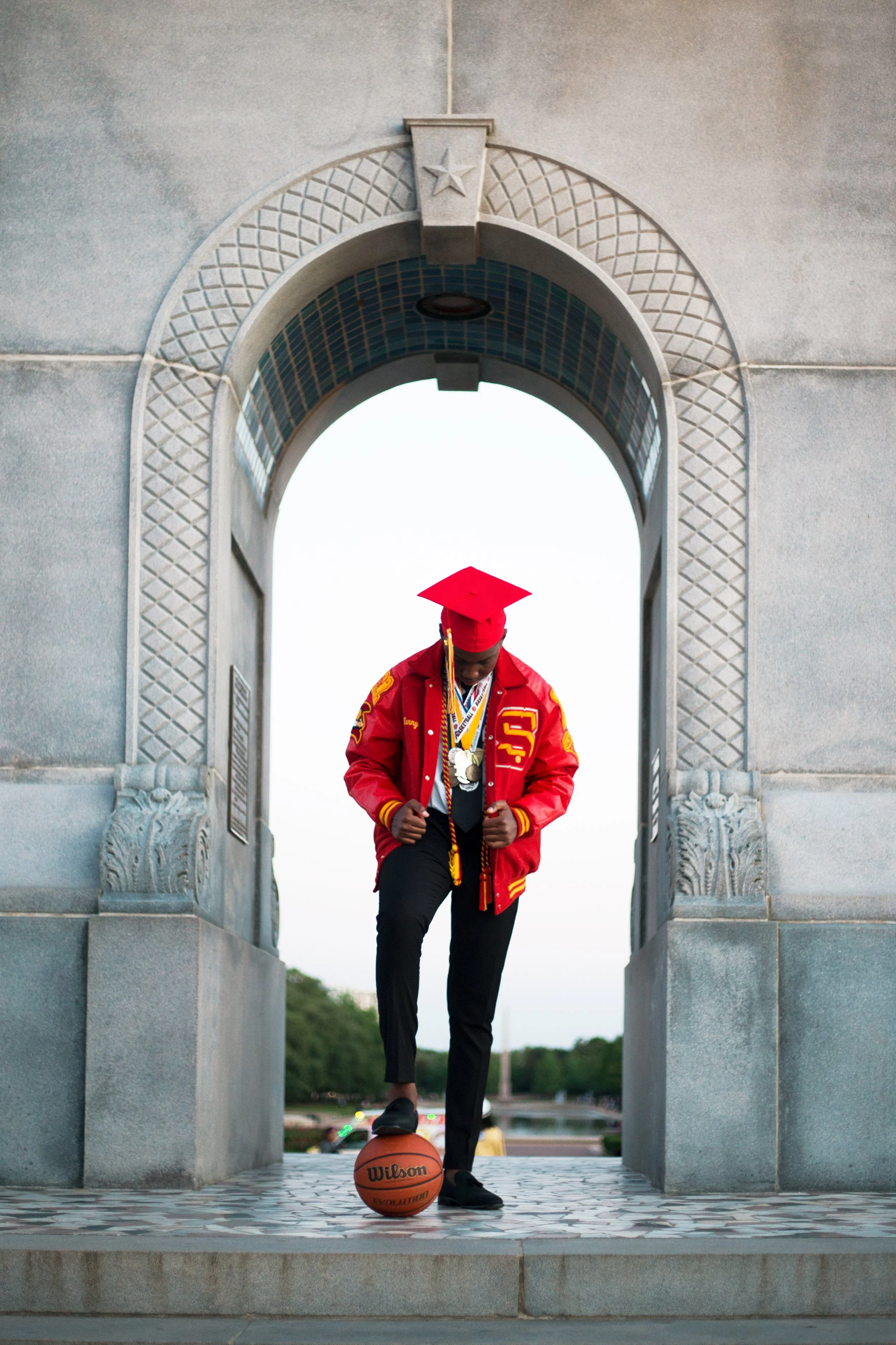 A young man in a red graduation cap and gown jacket with medals is playing basketball on a city street, with a stone archway in the background.