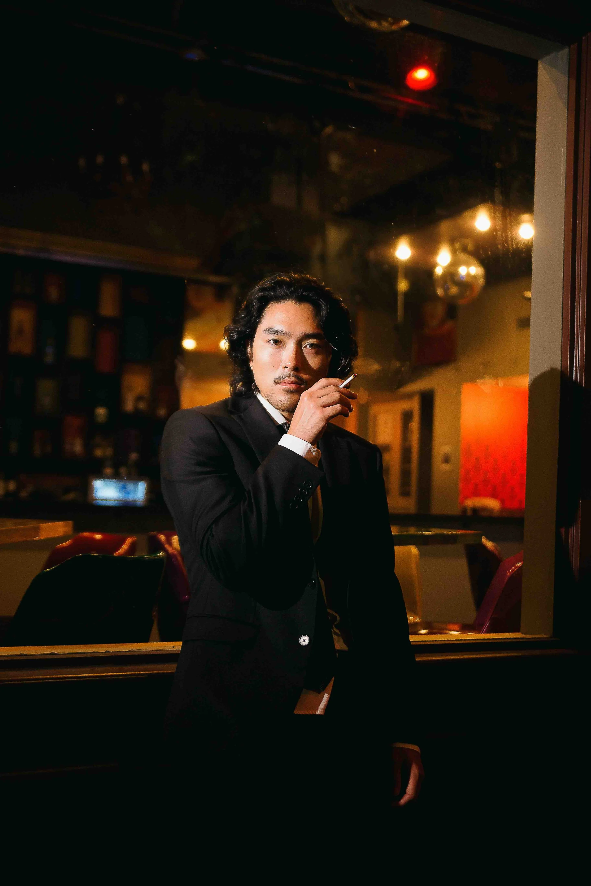 Houston cinematic portrait of a man in a black suit standing inside a dimly lit bar, holding a cigarette near his mouth and looking directly at the camera