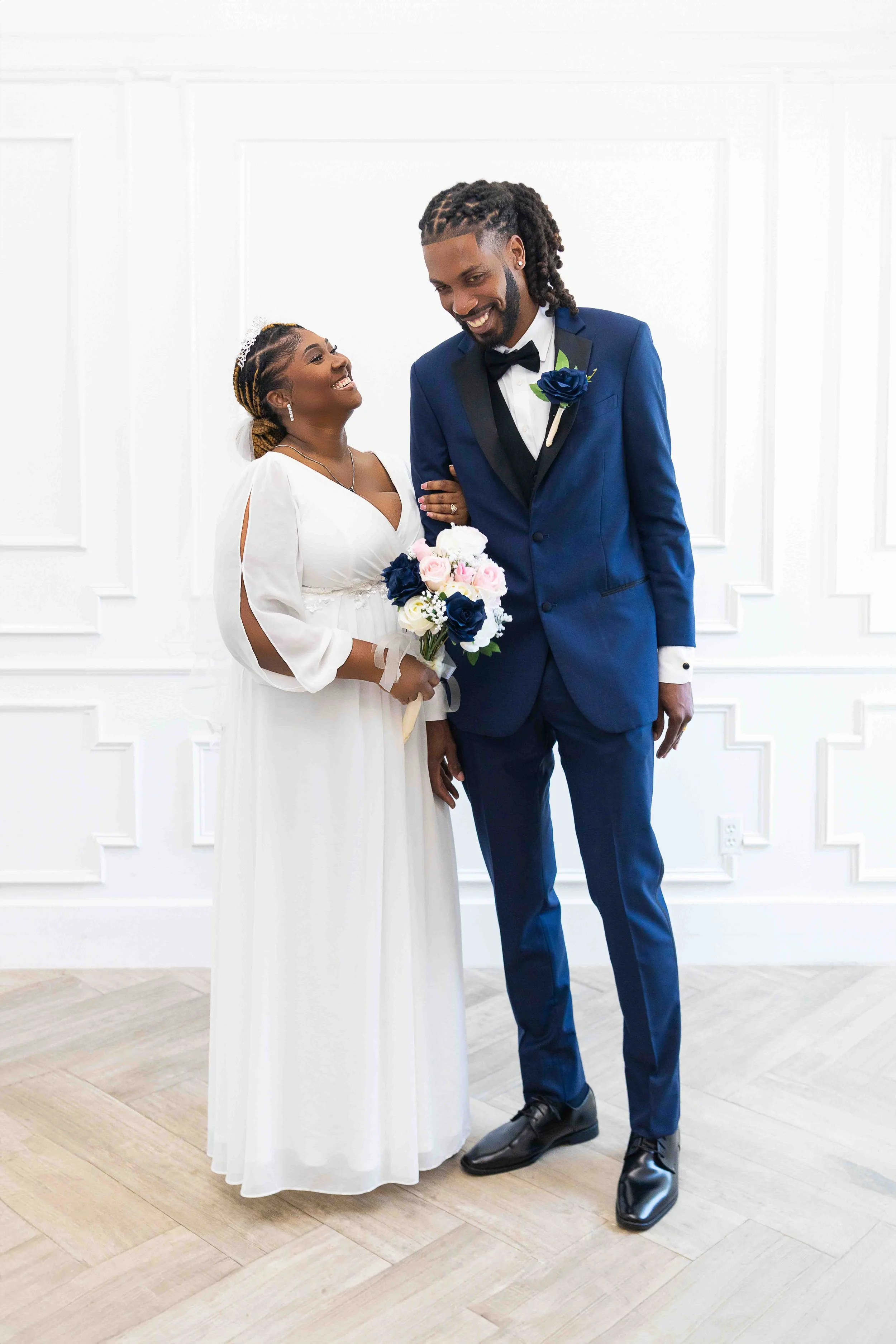 Elegant wedding portrait in Houston, Texas by Lev’s Photography featuring a smiling bride and groom looking at each other, with the bride holding a bouquet of pink, white, and dark blue flowers and the groom wearing a dark blue tuxedo with a black bo