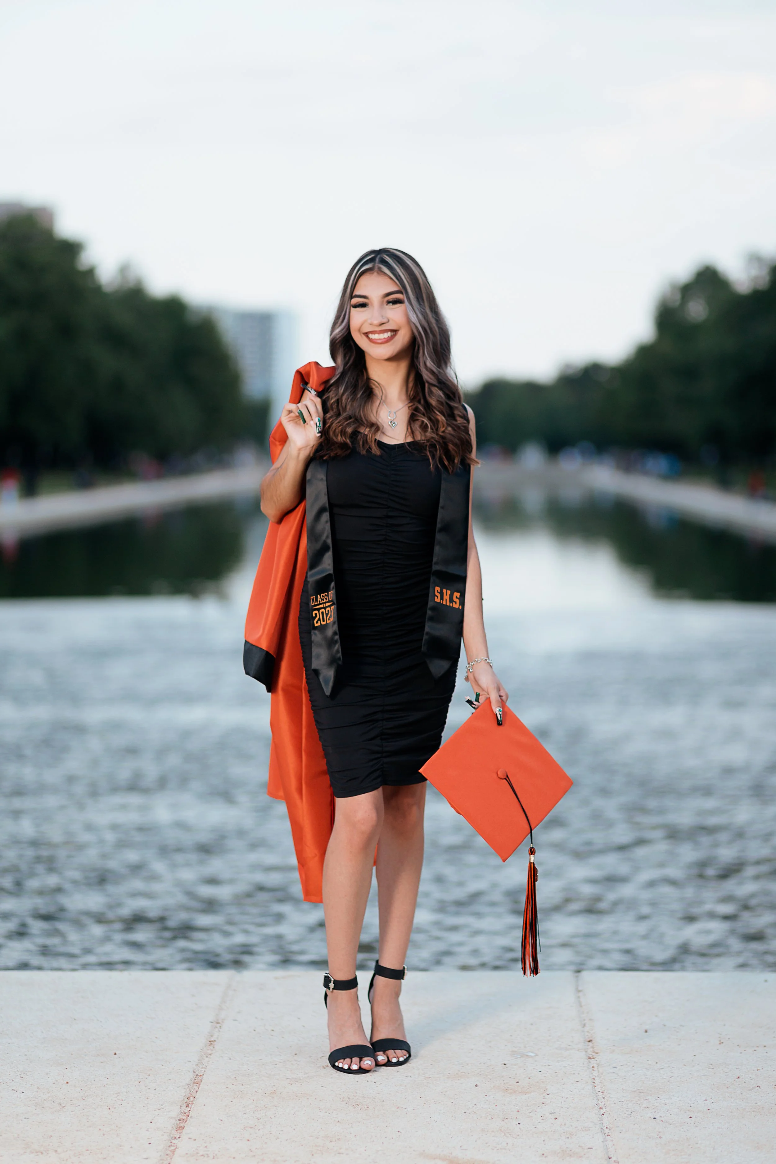 A young woman in a black dress and heels standing outdoors by a waterway, holding an orange graduation cap and wearing a graduation gown over her shoulders.