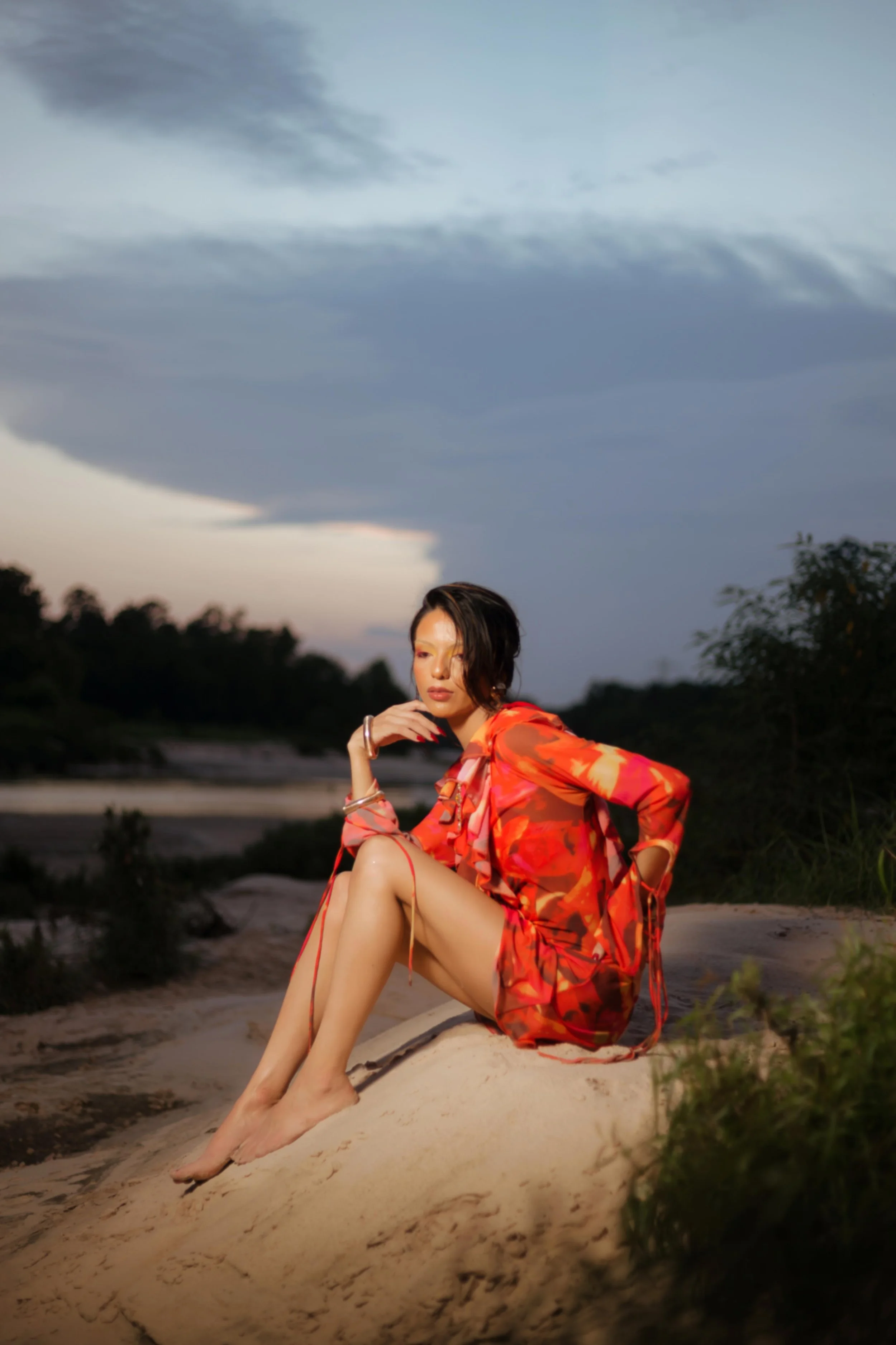 Humble, Texas editorial portrait by Lev's Photography featuring a woman in a vibrant red and orange dress sitting on a sand dune at dusk, captured with moody natural light and dramatic clouds, creative fashion-inspired outdoor portrait photography in