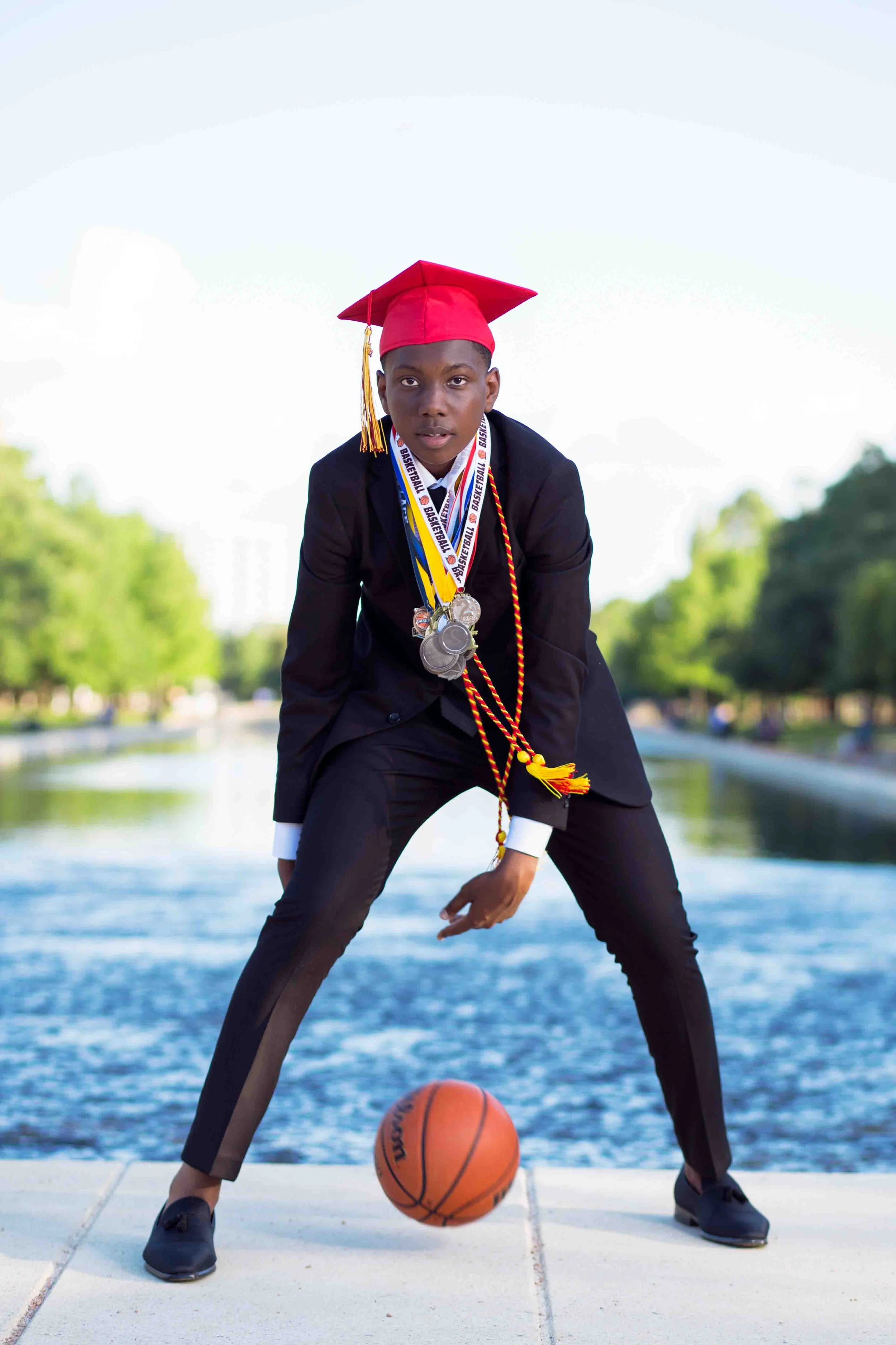 Graduation portrait at Hermann Park in Houston featuring a young man in a black suit and red graduation cap with tassel, wearing medals and honor cords, standing outdoors on a walkway by a canal with a basketball at his feet, leaning slightly forward