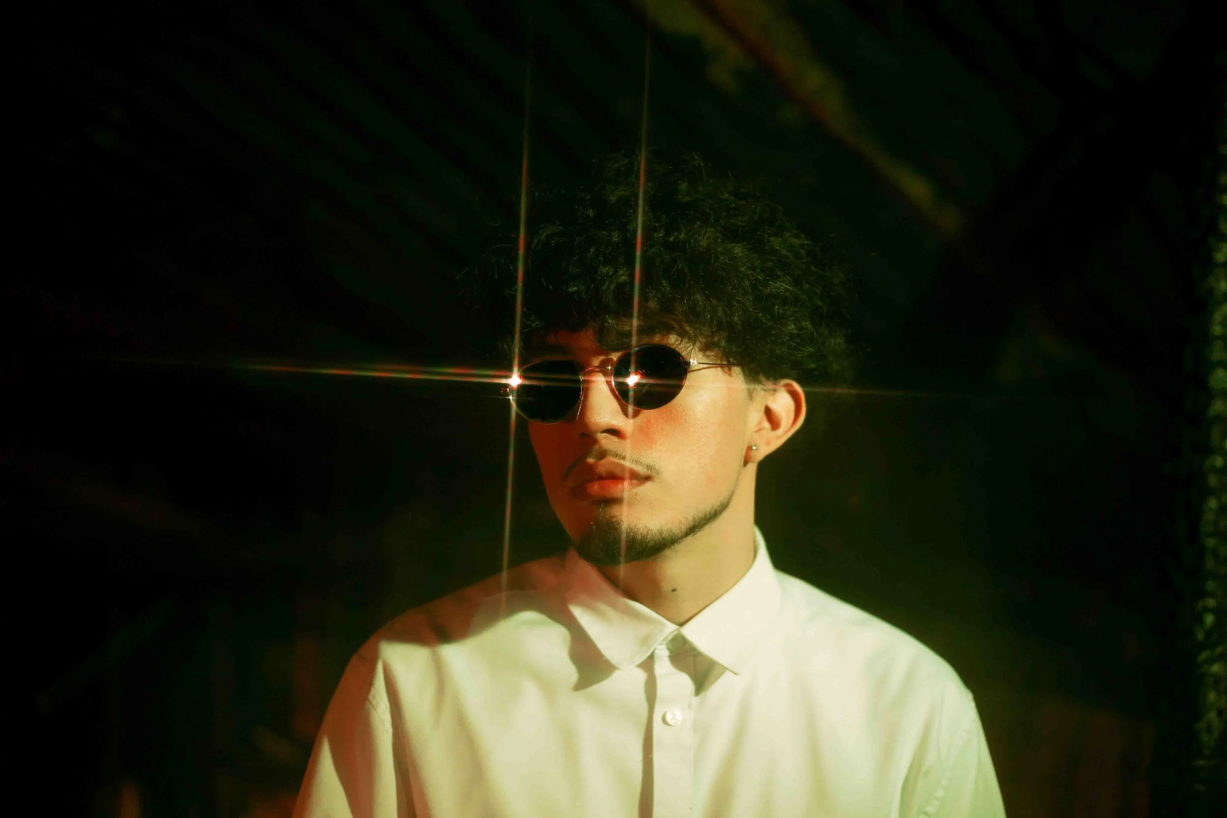 Houston portrait of a young man with curly hair and sunglasses standing in a dark setting with starburst light reflections on the lenses
