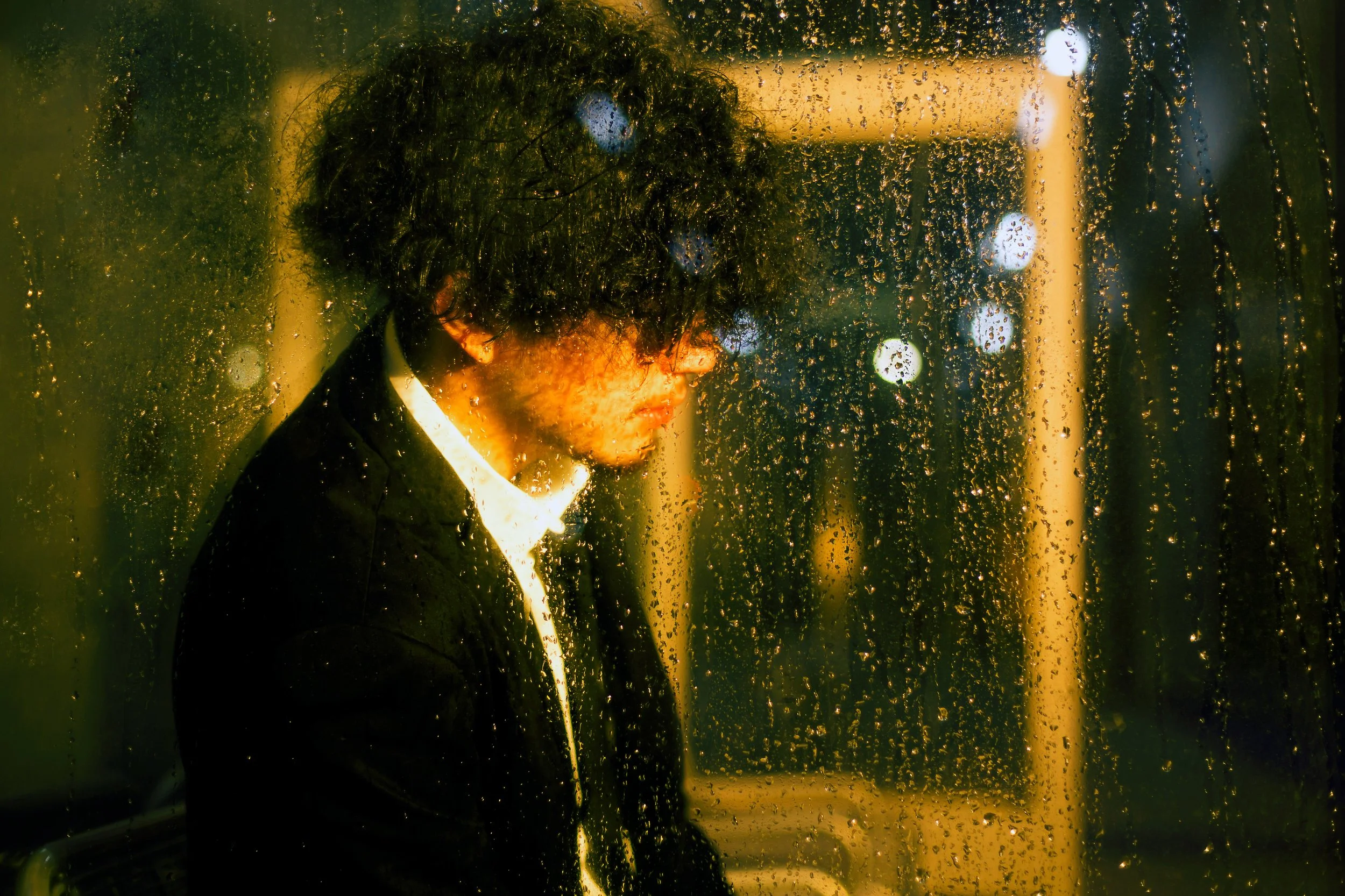 A person with curly black hair and glasses wearing a black suit and white shirt, seen through a rain-covered glass window at night, with blurred city lights in the background.