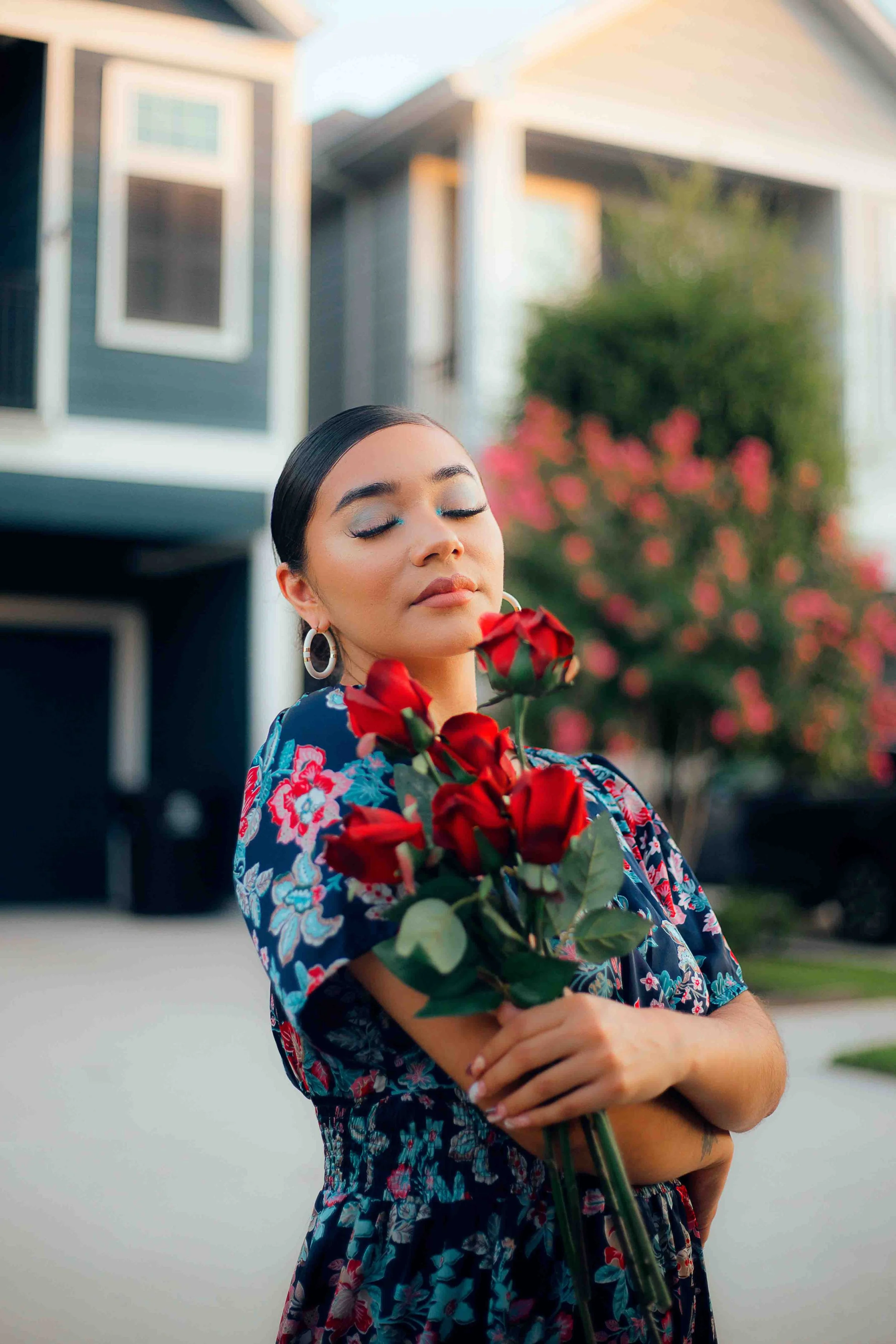 Houston portrait of a woman with closed eyes holding a bouquet of red roses in front of her chest, standing outdoors in front of a suburban house with a pink-flowered tree.