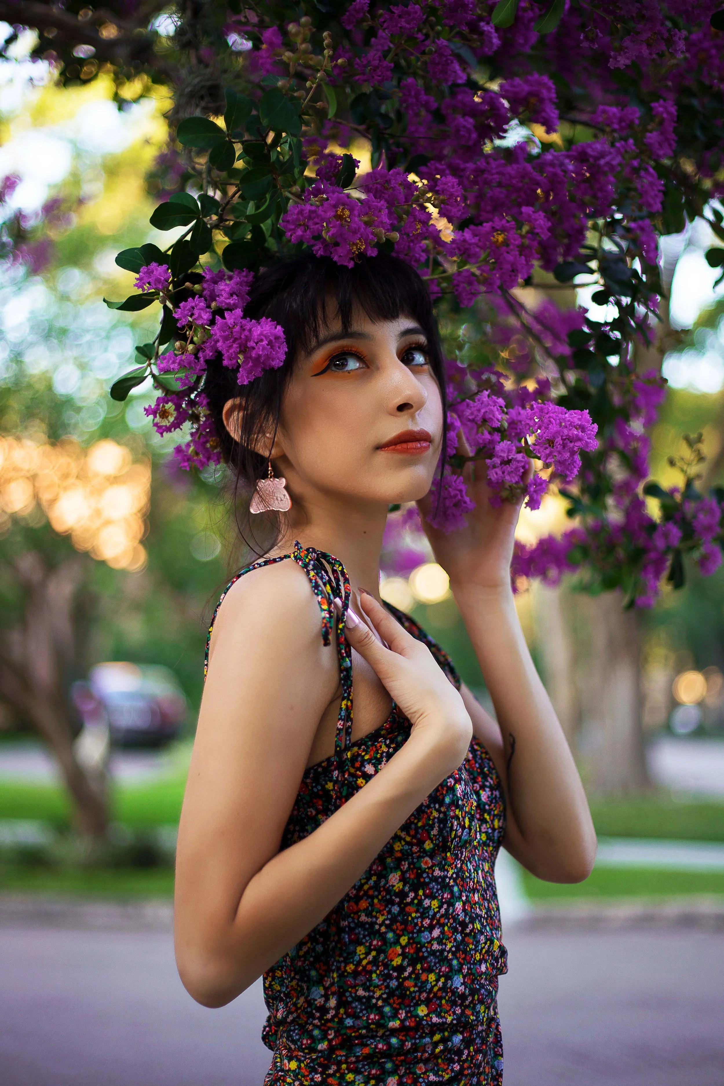 A woman wearing a floral dress with colorful small flowers, standing outdoors in a park, holding purple bougainvillea flowers above her head, with a blurred background of trees and a sidewalk, during sunset.