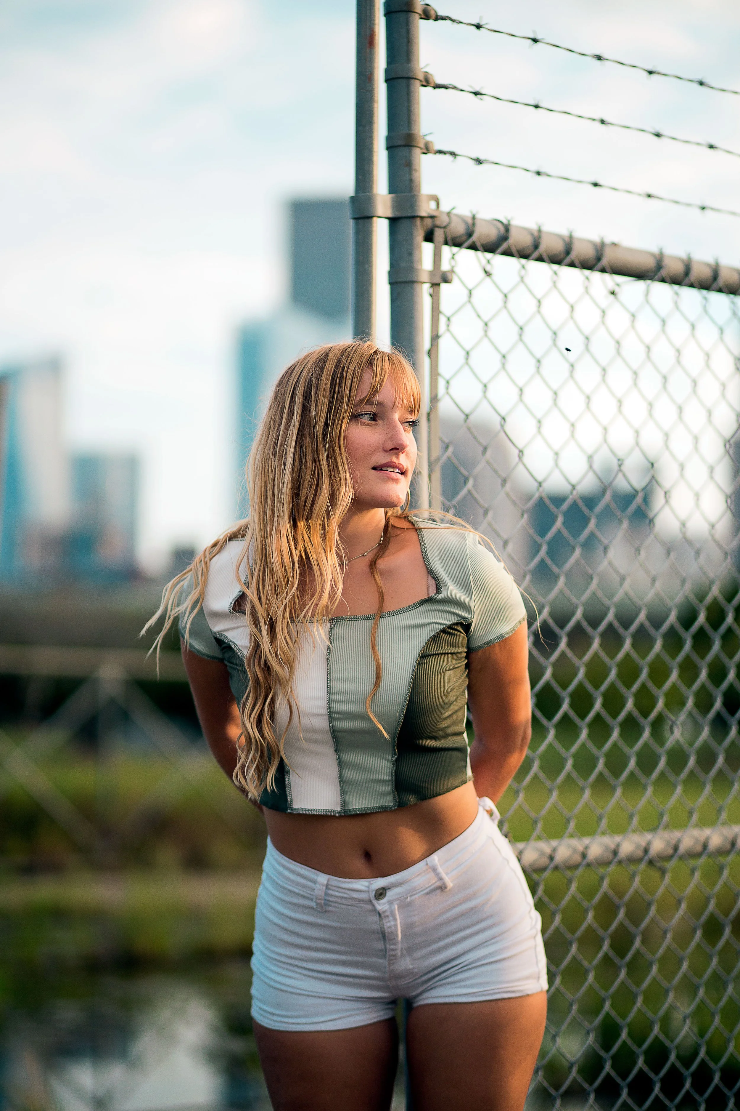 A young woman with long wavy red hair leaning against a chain-link fence, in an urban setting with city buildings in the background during daylight.