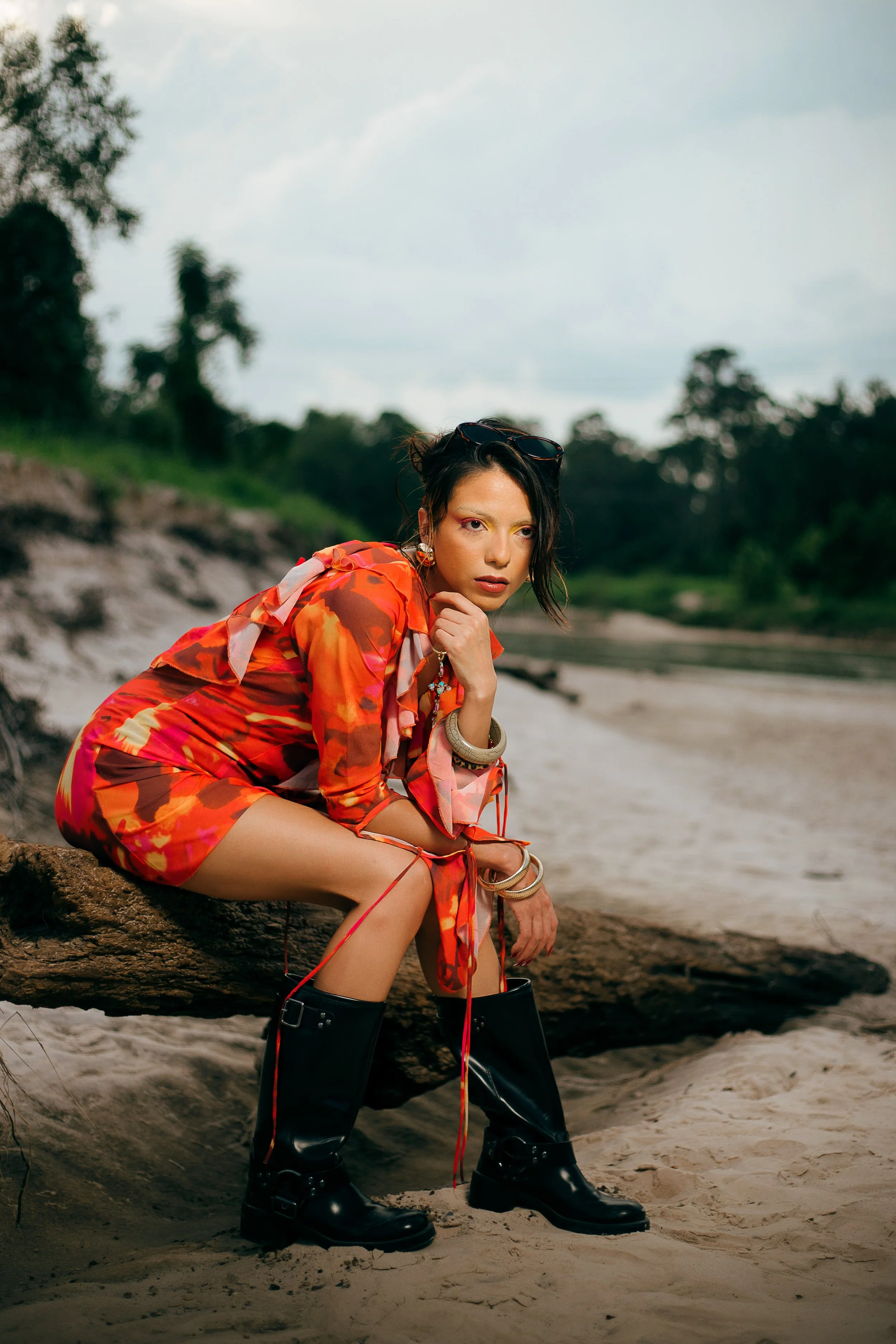 A woman dressed in a colorful red and orange patterned outfit, sitting on a log on a sandy beach with greenery and cloudy sky in the background.