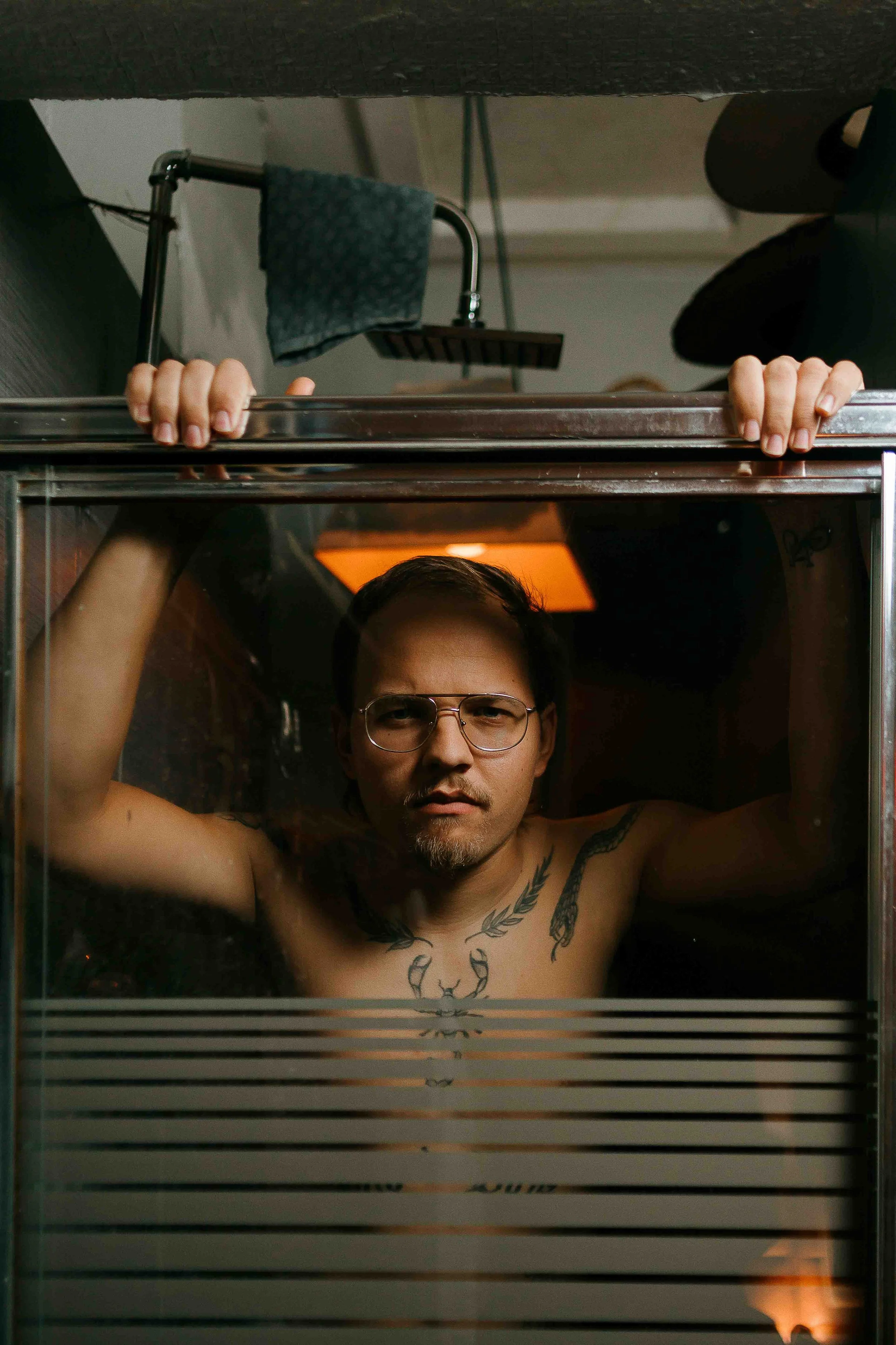 Cinematic editorial portrait of a tattooed man with glasses gripping a glass enclosure in a dimly lit room with a warm orange ceiling light, movie-style lighting