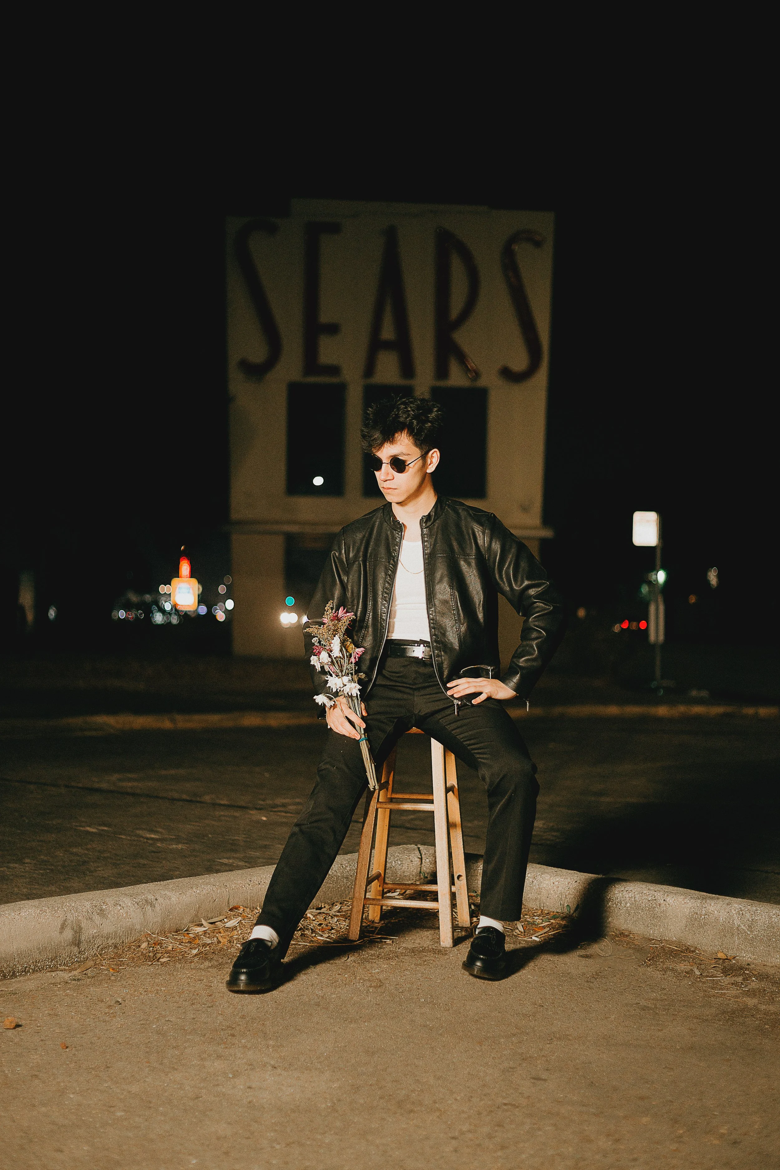 A person sitting on a stool in a parking lot at night, holding a bouquet of flowers, wearing sunglasses, a leather jacket, black pants, and black shoes, with a large sign reading 'SEARS' illuminated in the background.