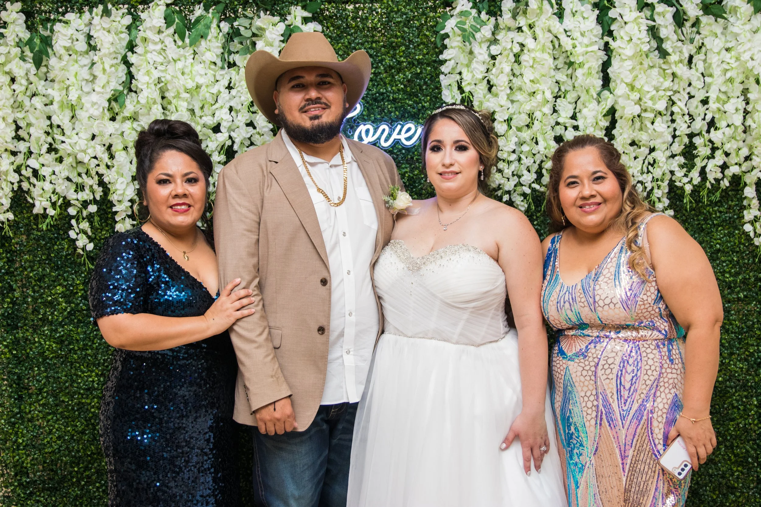 A group of five people, including a bride and groom, standing in front of a floral backdrop at a wedding celebration. The bride is wearing a white strapless wedding gown, and the groom is dressed in a beige suit with a cowboy hat.