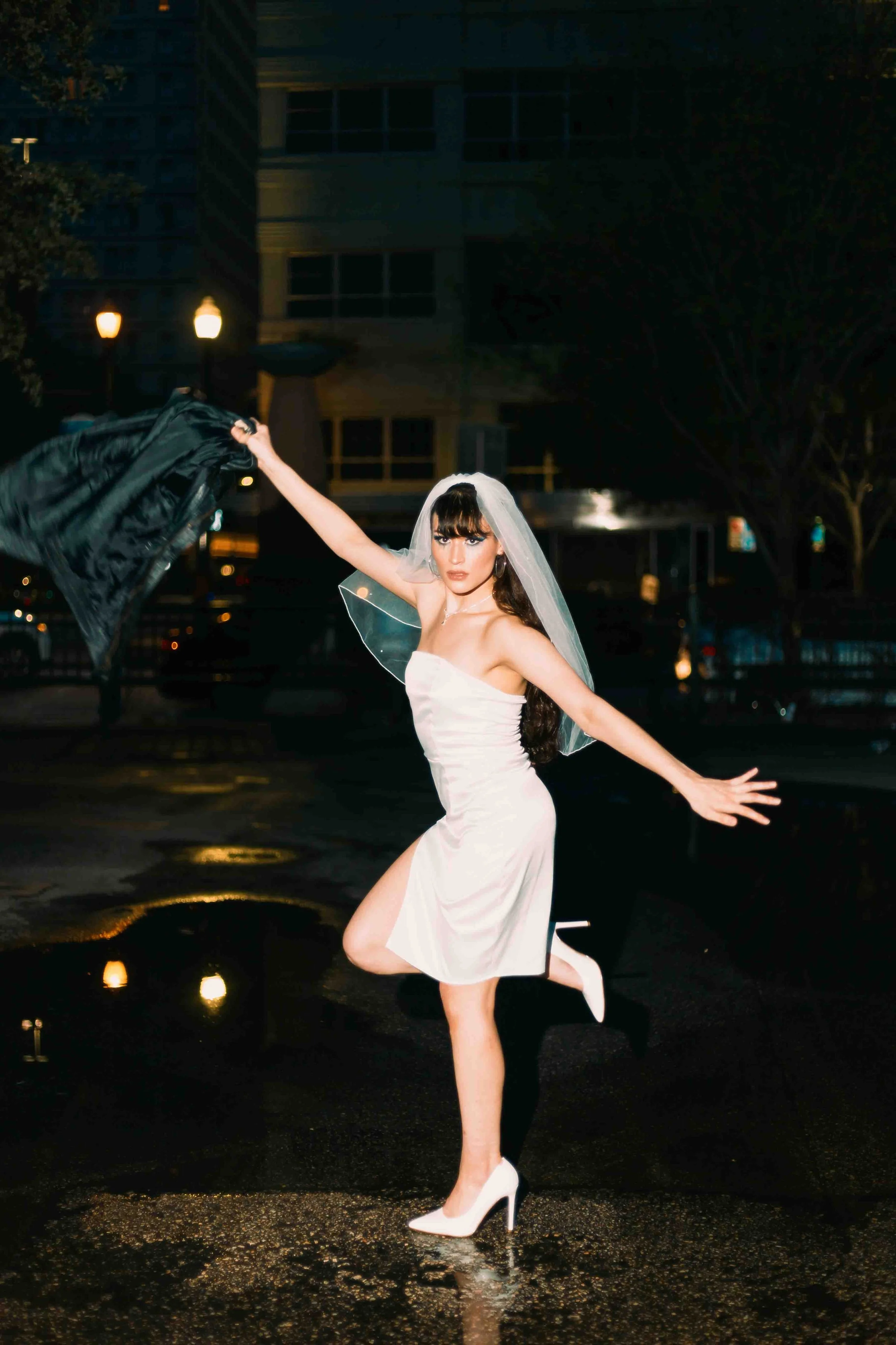 Houston editorial portrait of a woman in a white dress and high heels dancing outdoors at night, holding a black jacket with cityscape and streetlights in the background