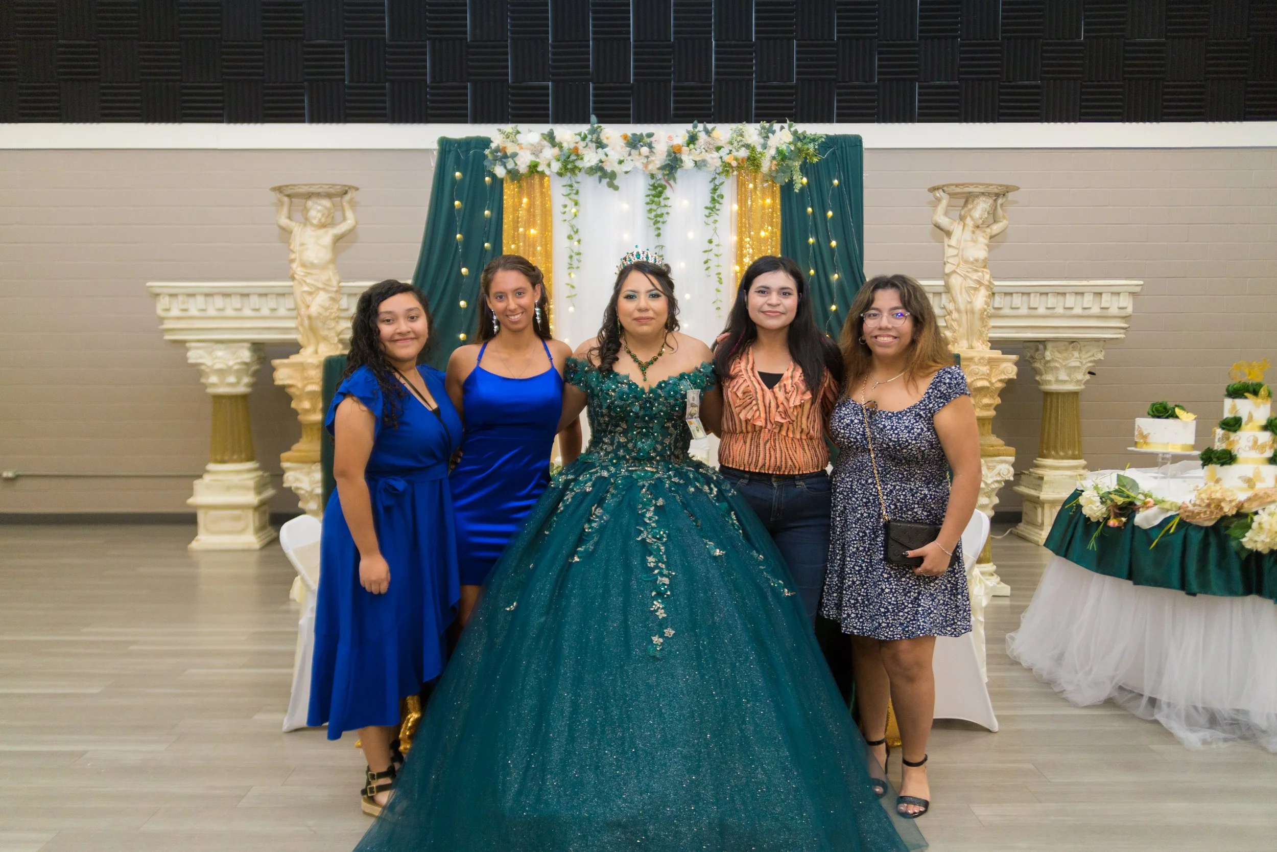 Group of five women, including a girl in an elaborate emerald dress with a tiara, celebrating at a formal occasion with decorated backdrop and desserts.