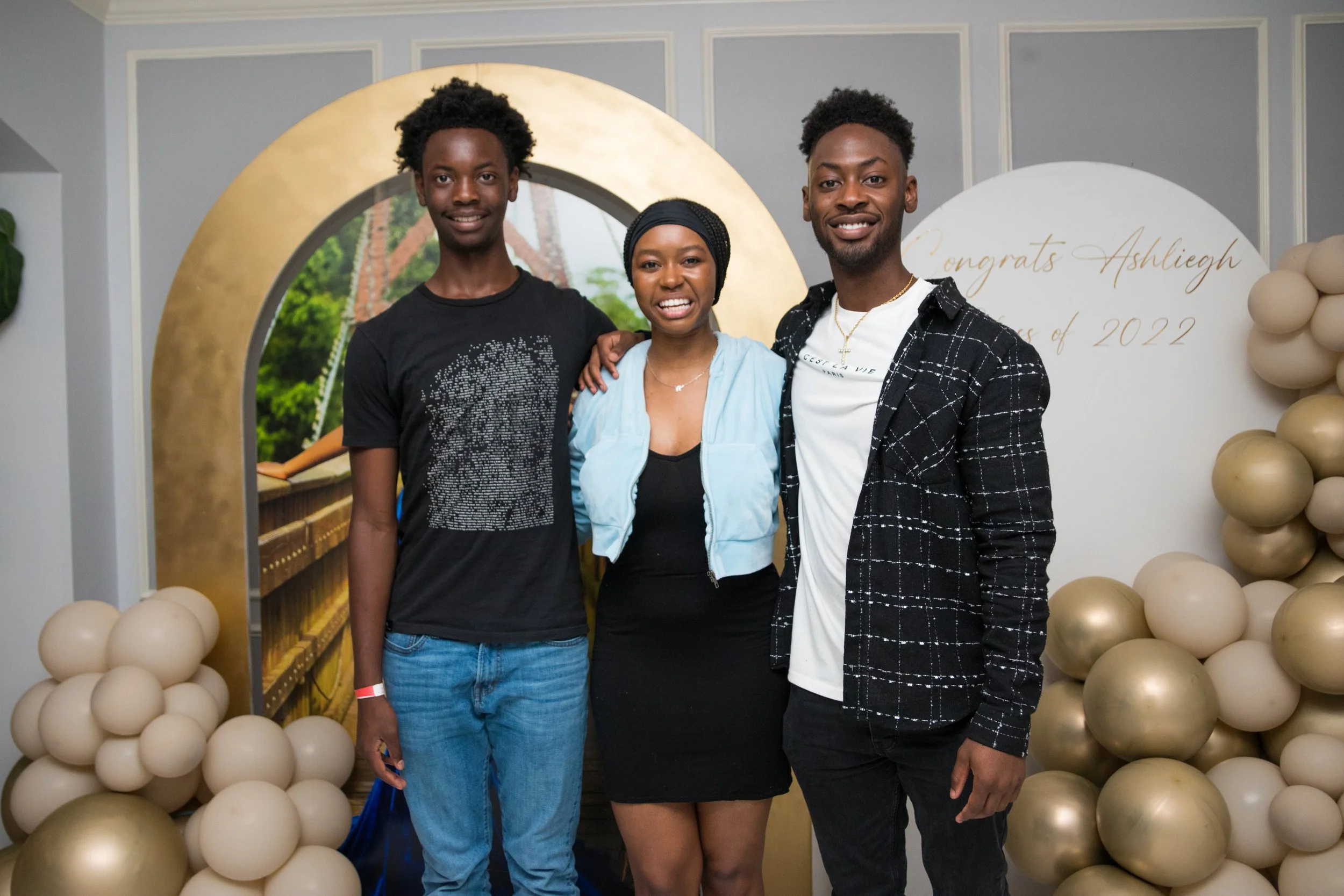 Three young adults, two men and one woman, are standing together at a celebration with a decorative backdrop and balloons. The woman is in the middle, wearing a turquoise jacket and black dress, while the men are on either side, one in a black T-shir