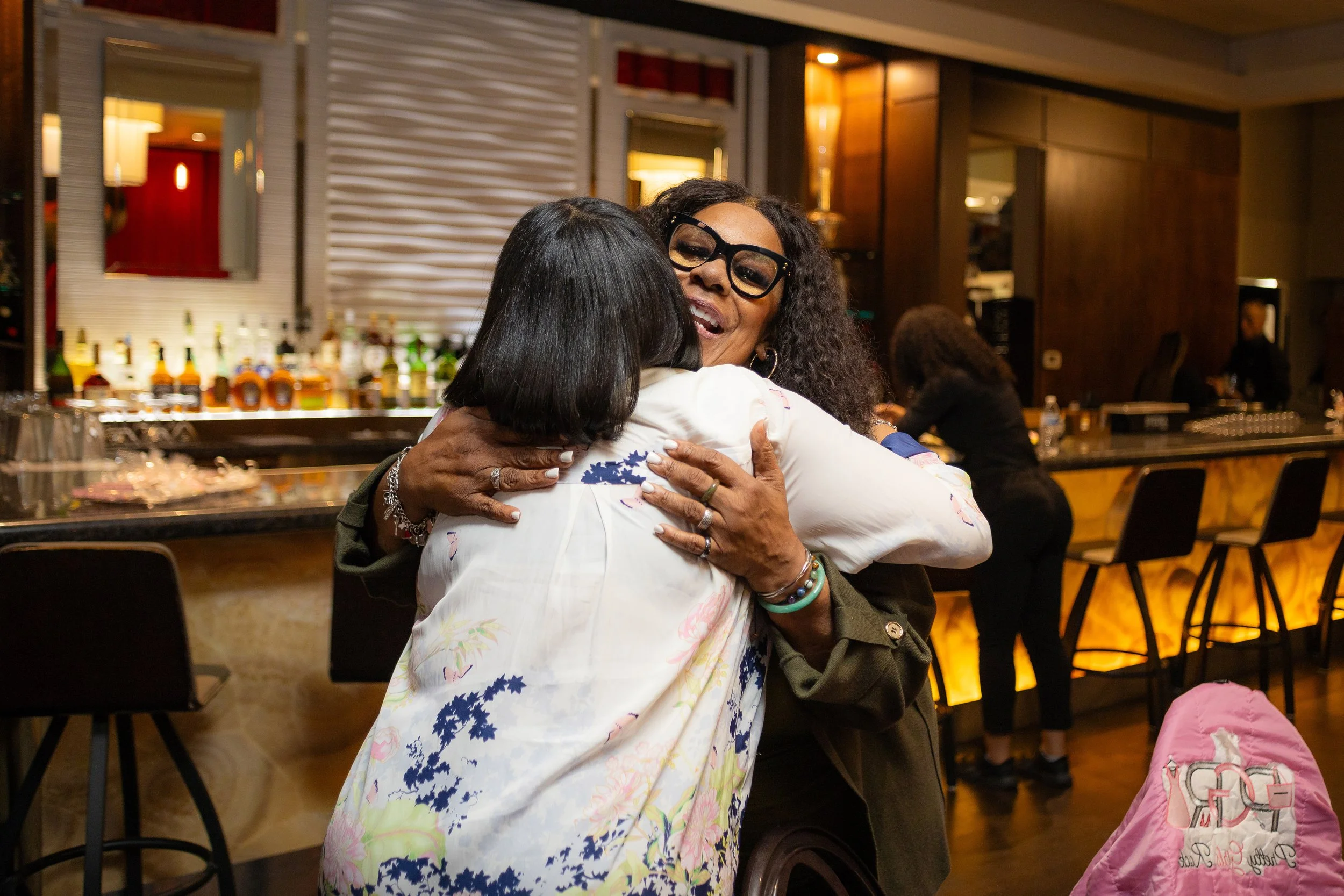 Two women hugging warmly in a bar or restaurant setting with a bartender and bar in the background.