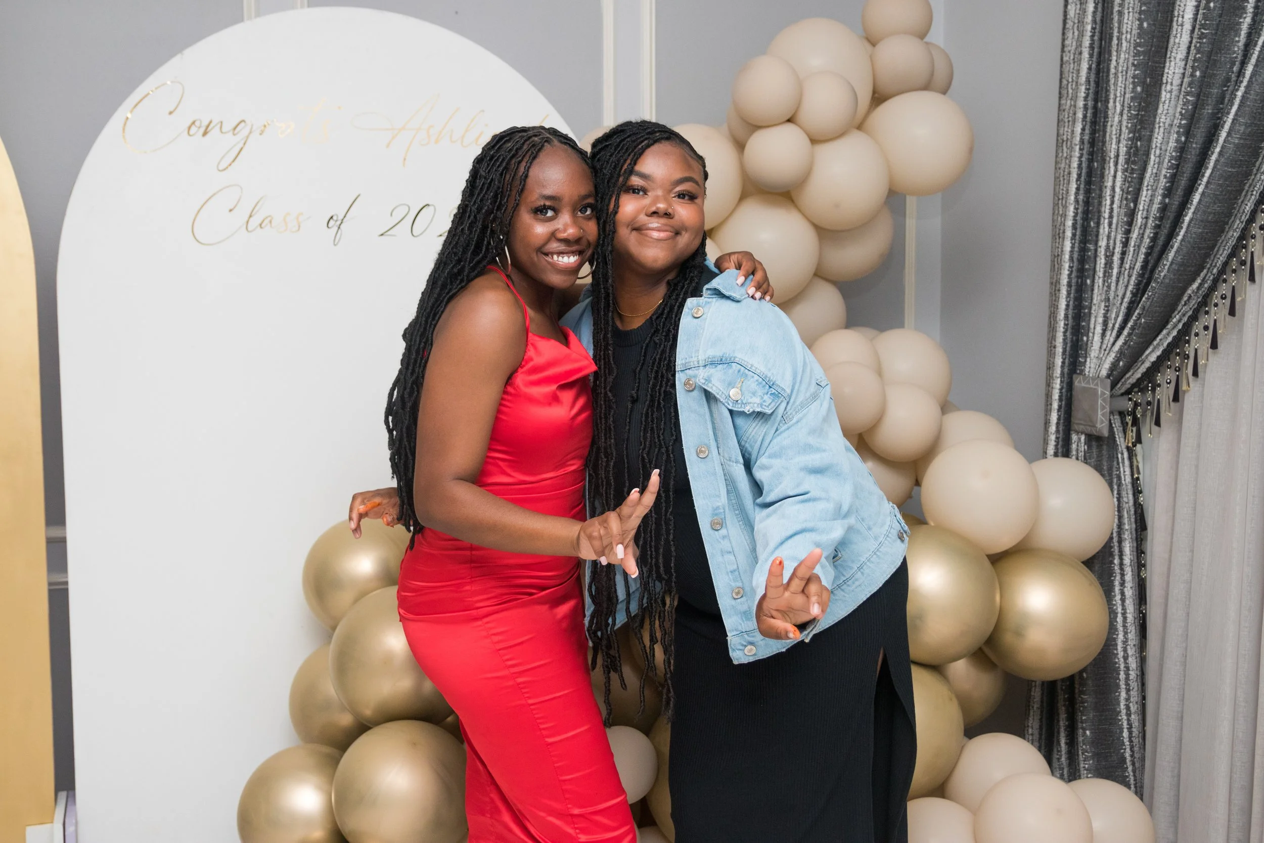 Two young women are smiling and posing together at a celebration. One is wearing a red dress, and the other is wearing a black outfit with a denim jacket. They are standing in front of a backdrop with cream-colored balloons and a white display board