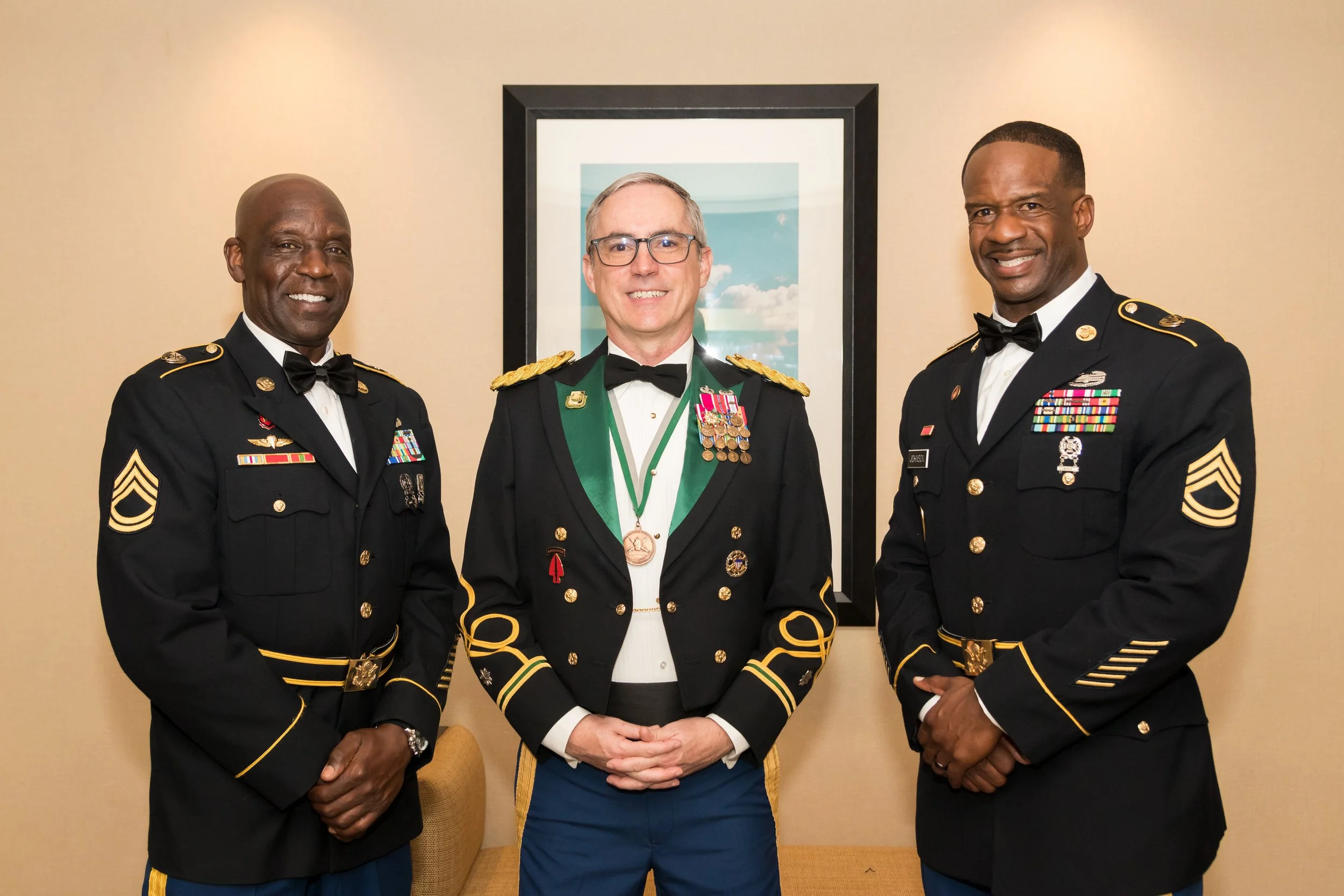 Three military officers in dress uniform standing together indoors, smiling, with a framed sky scene in the background.