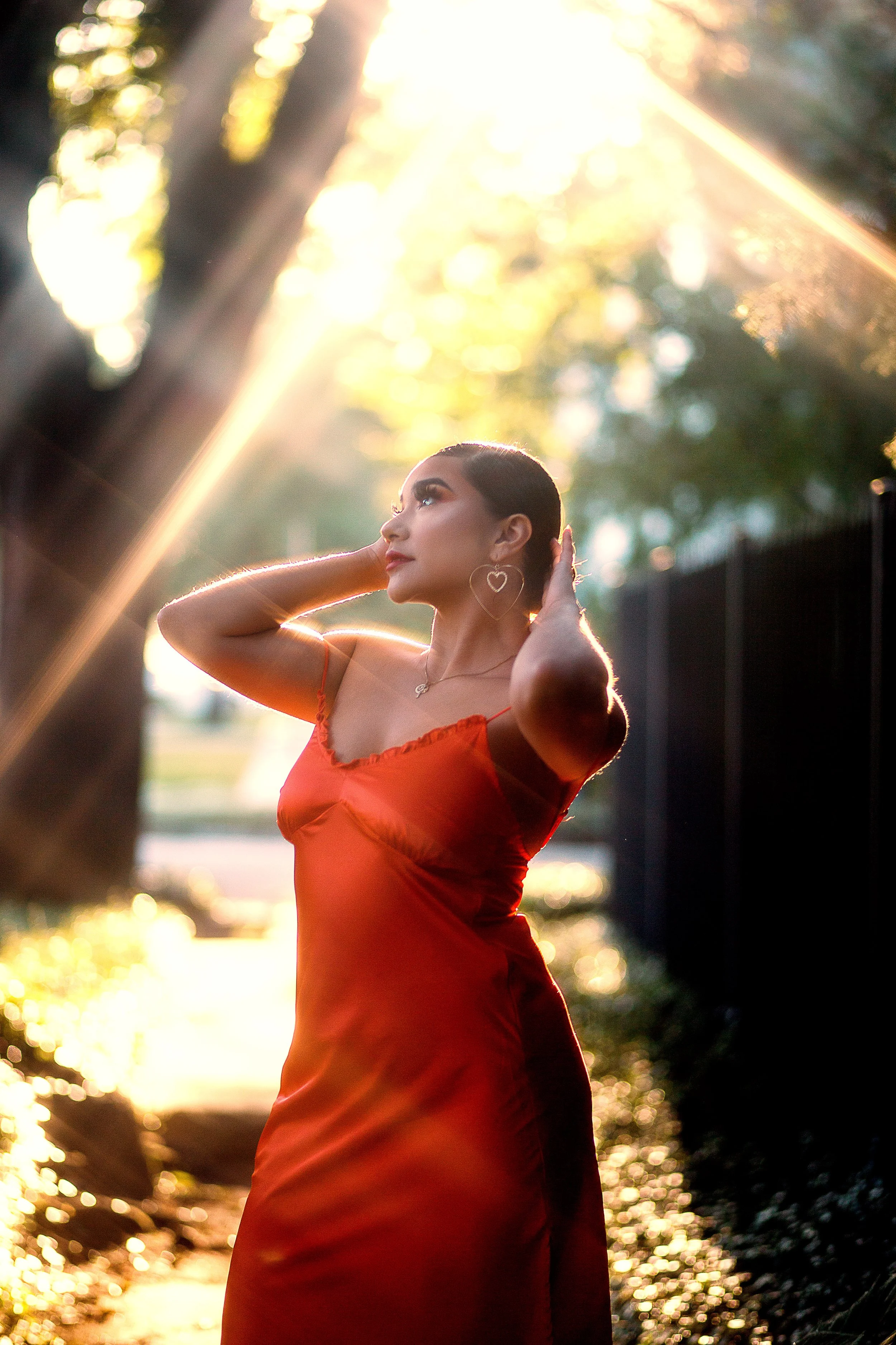 A woman in a red dress standing outdoors during sunset, with sunlight filtering through trees behind her, posing with hands behind her head and looking to the side.