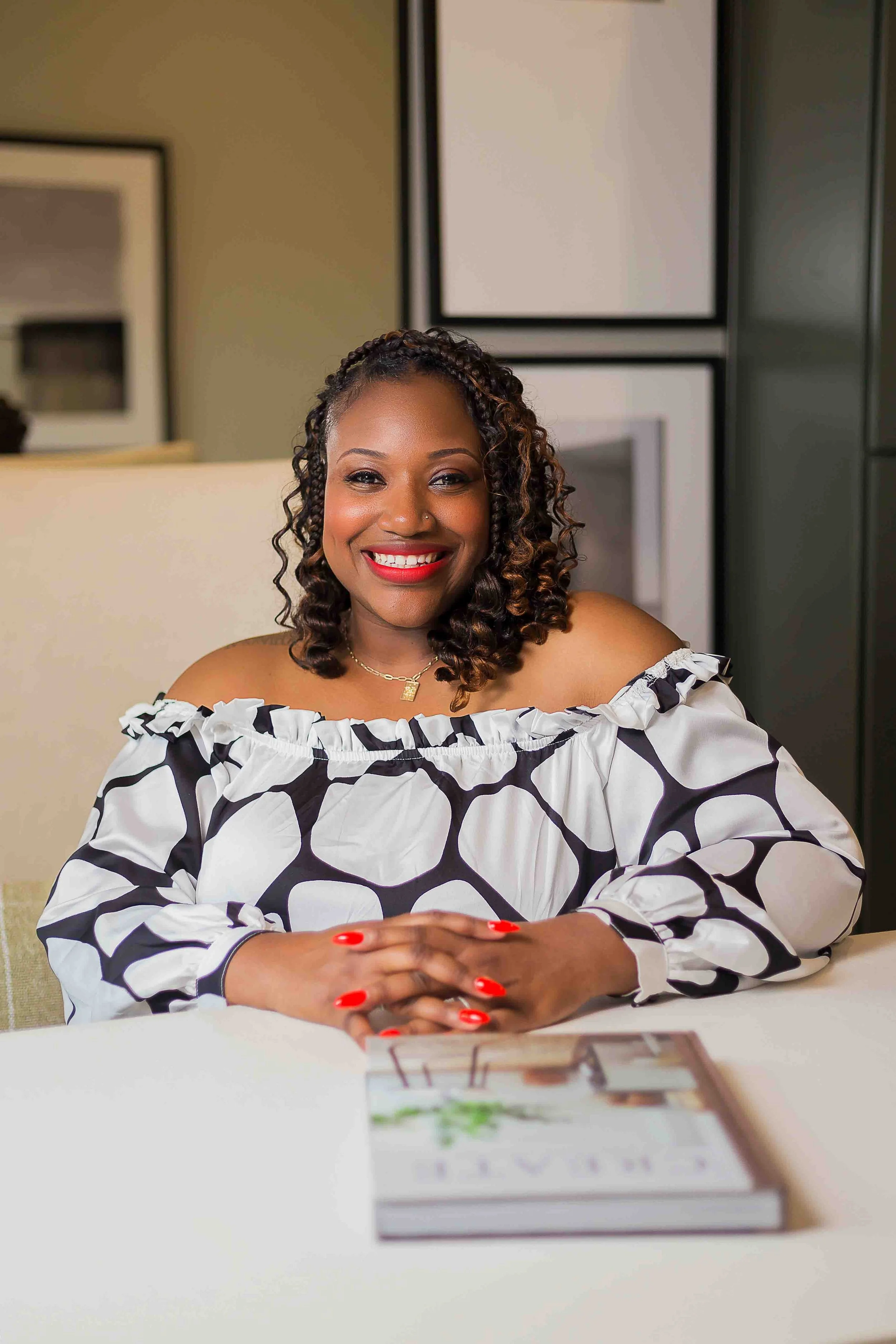 Houston realtor branding session by Lev’s Photography featuring a smiling woman with curly hair and red lipstick seated at a table with a magazine in a modern interior setting