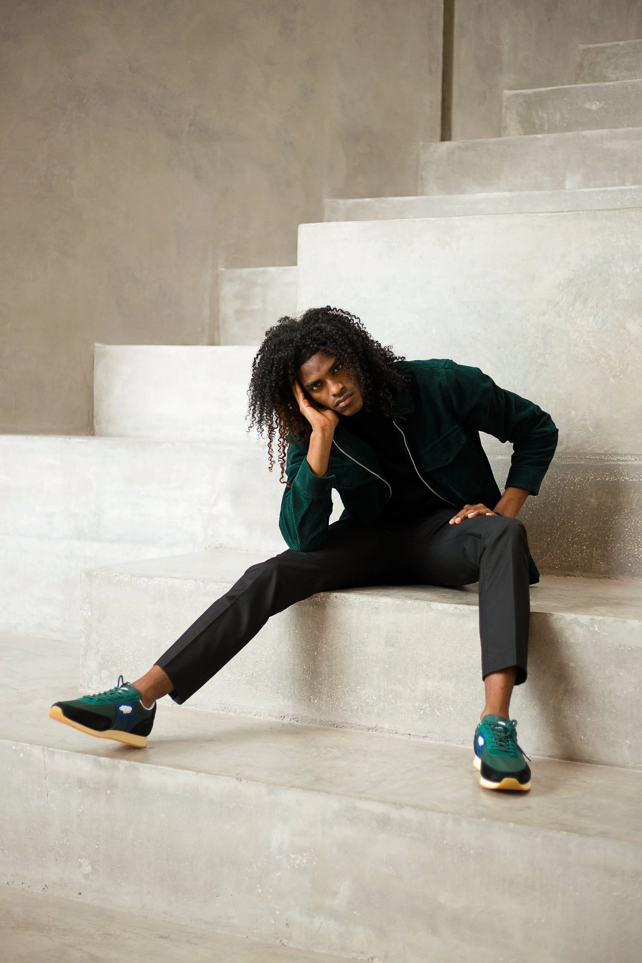 A woman with curly hair sits on concrete stairs, leaning her head on her hand, wearing a dark jacket, black pants, and green sneakers.