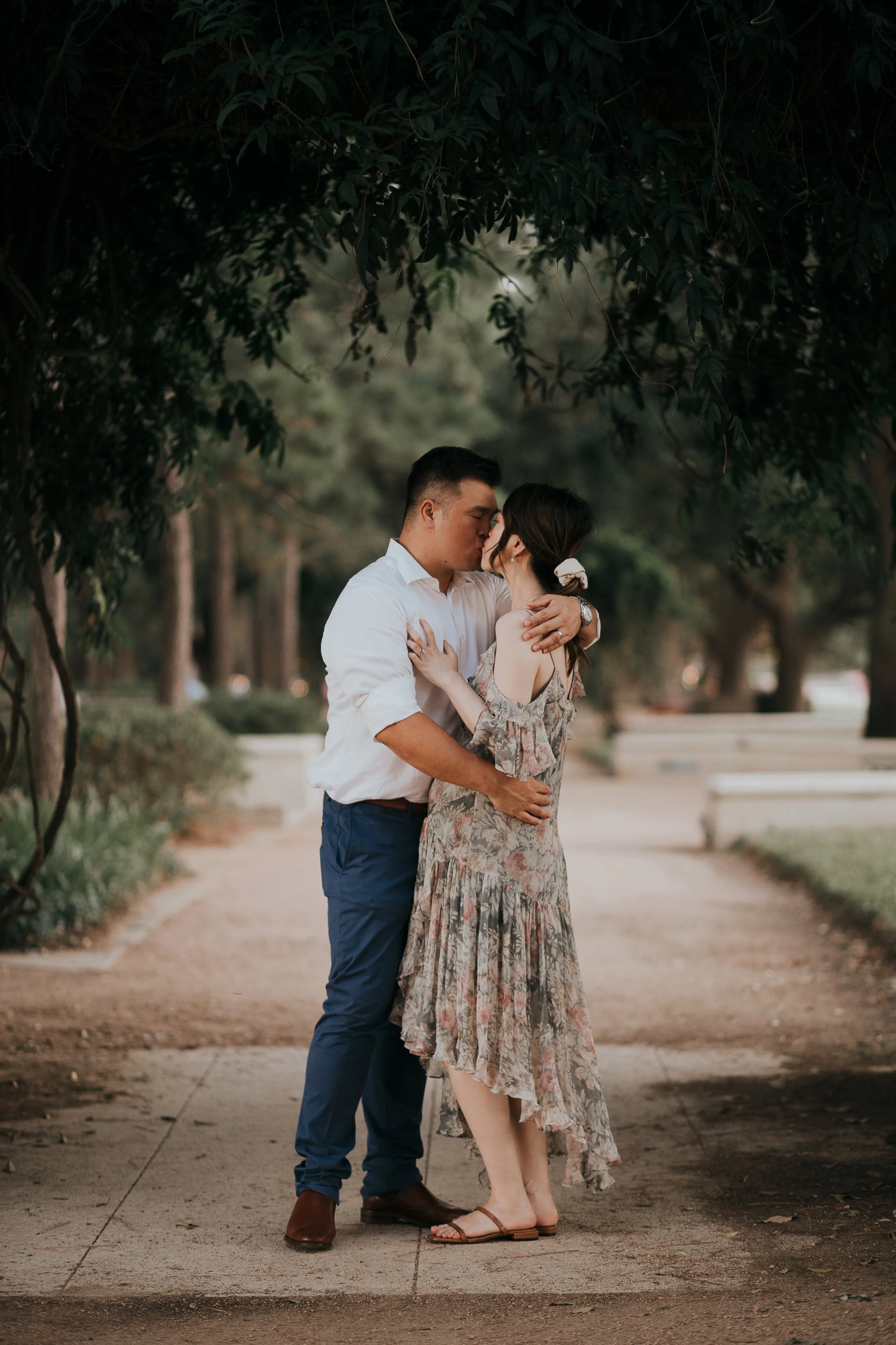 A romantic couple sharing a kiss in a park, standing close under a large tree with a pathway and greenery in the background.